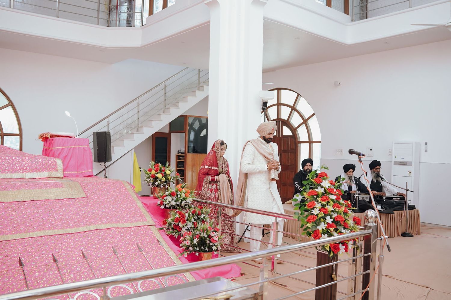 Sikh wedding ceremony inside a gurdwara with couple standing near the decorated Guru Granth Sahib stage.