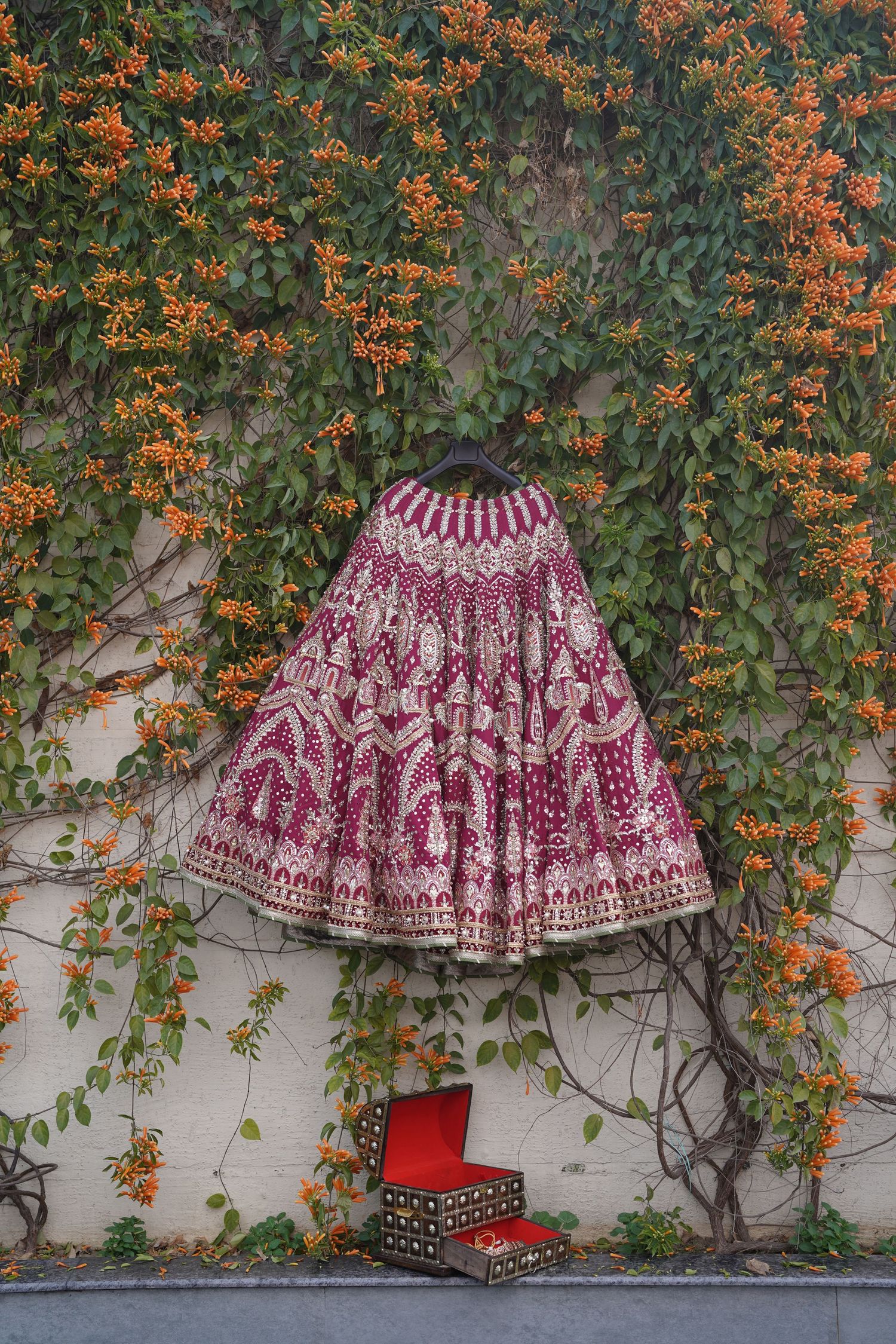 A heavily embroidered maroon and gold bridal lehenga displayed against a wall covered in orange flowering vines.
