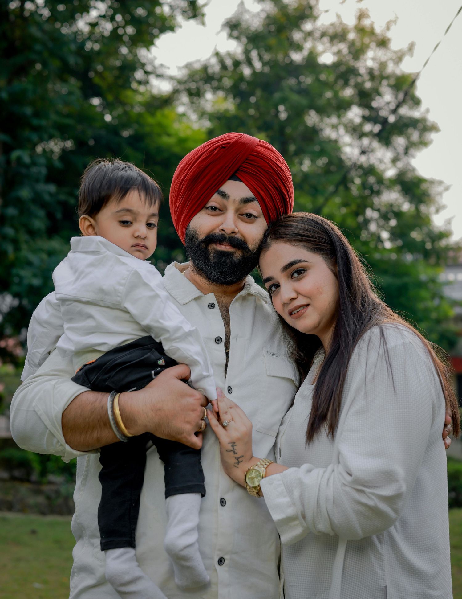 Sikh father in red turban holding toddler son while mother leans in for outdoor family portrait.