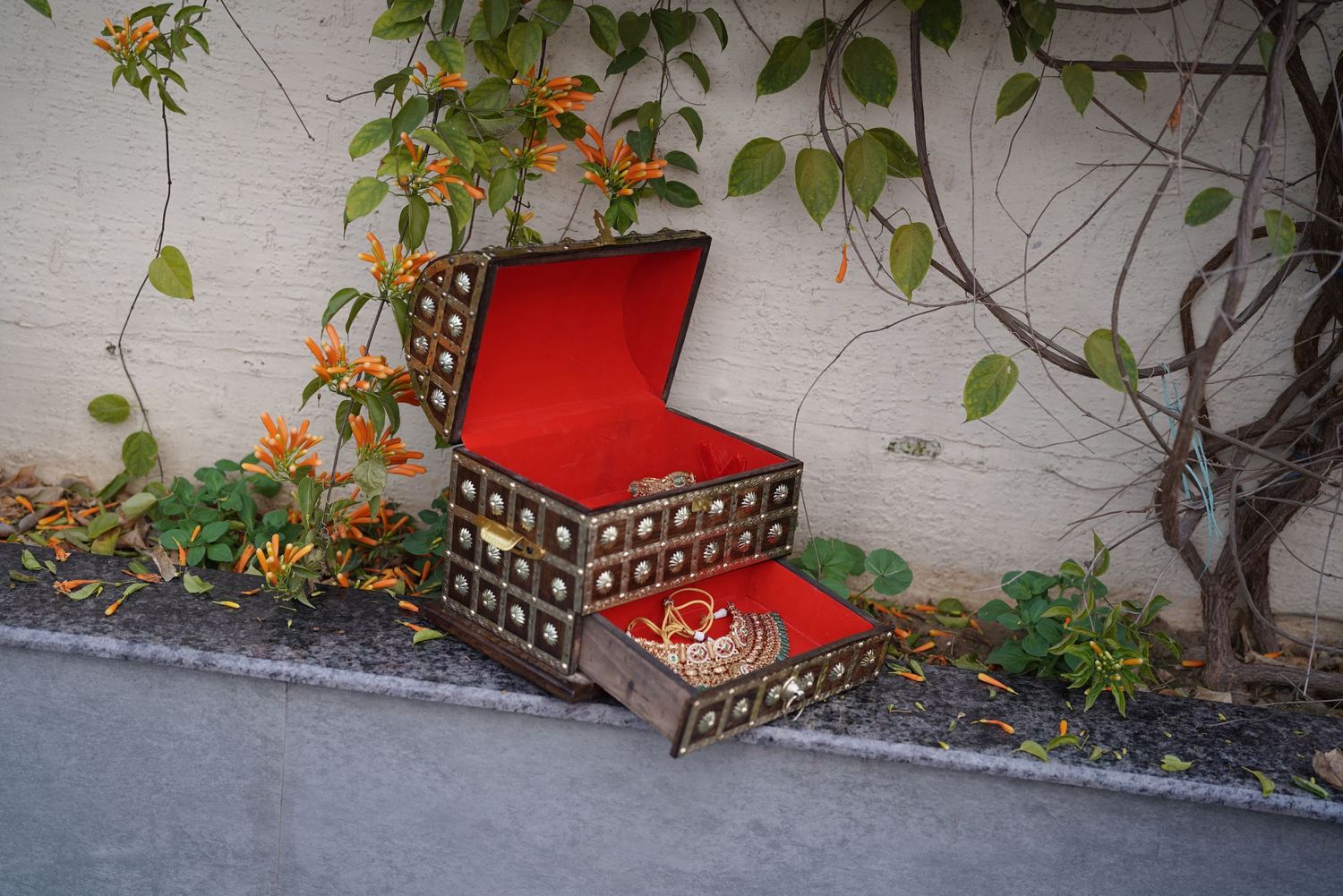 An ornate wooden jewelry box with red velvet interior, open and displayed outdoors near flowering vines.