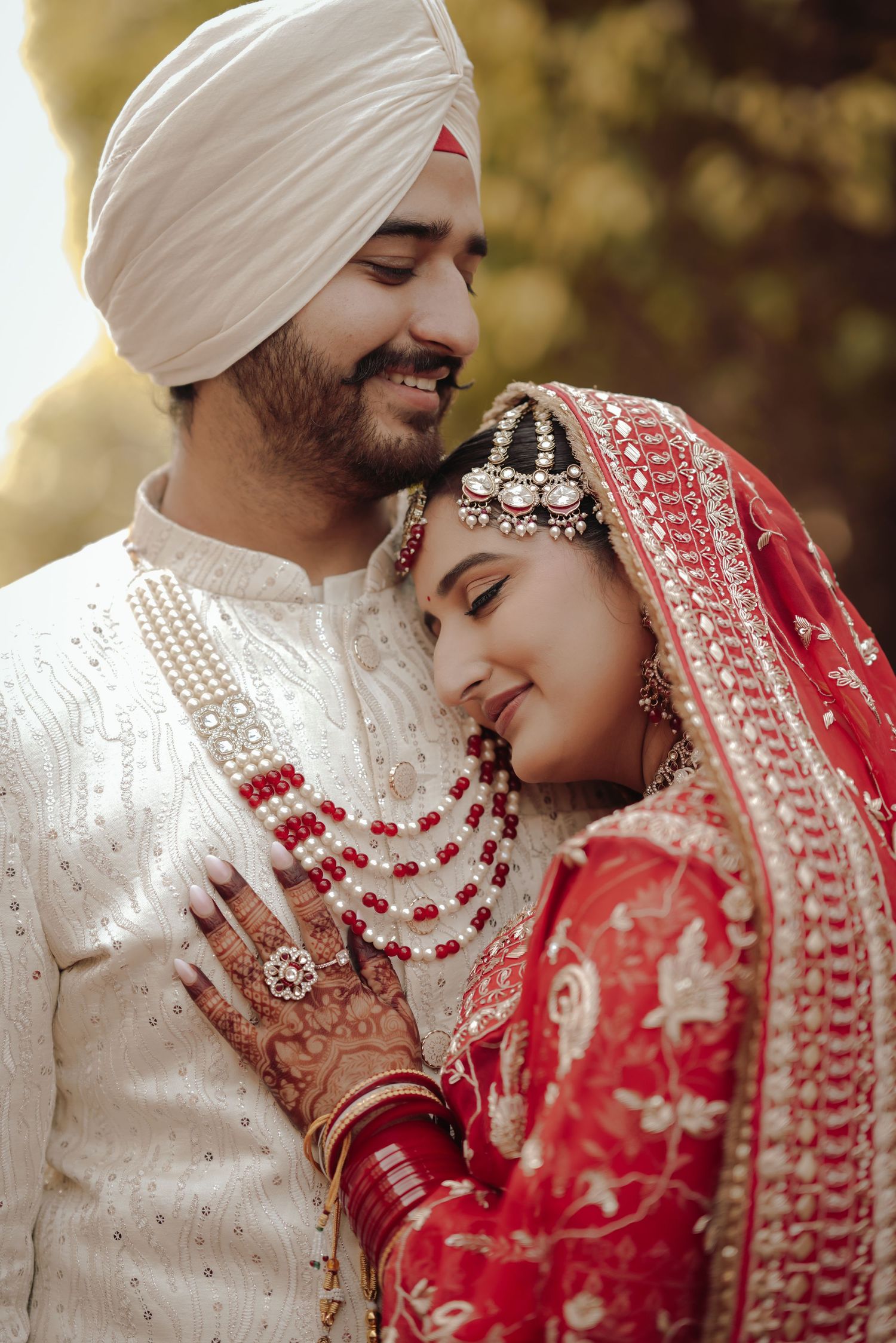 Sikh groom in white sherwani and turban tenderly embracing bride in red lehenga with pearl necklace.