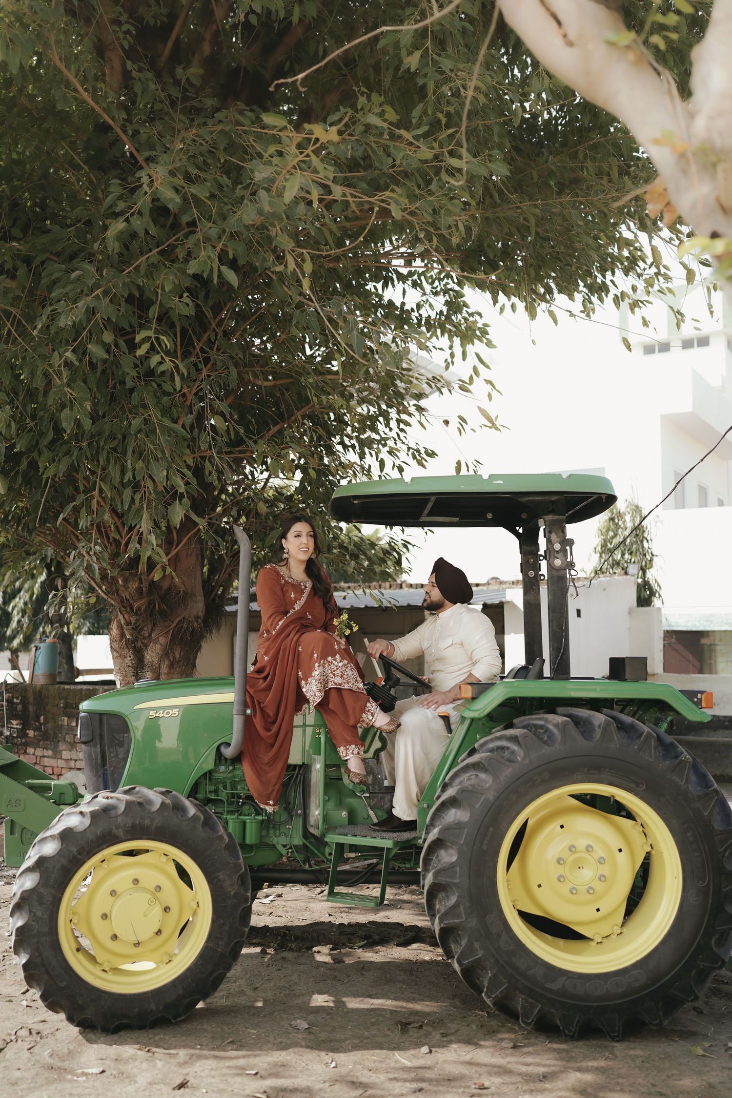Sikh couple in traditional outfits posing on a green John Deere tractor under trees.