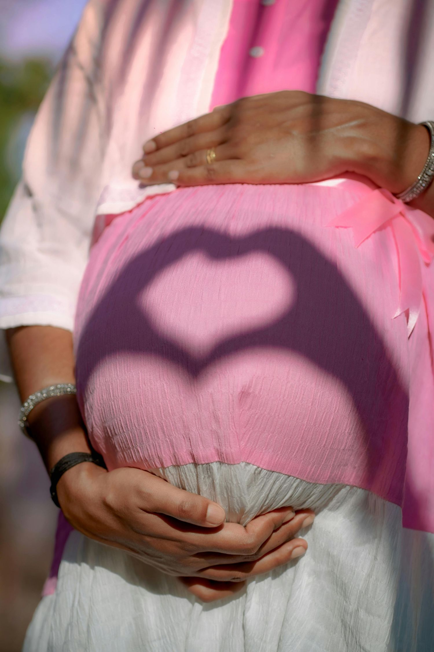 Heart shadow cast on pregnant belly with couple's hands forming love symbol in pink light.
