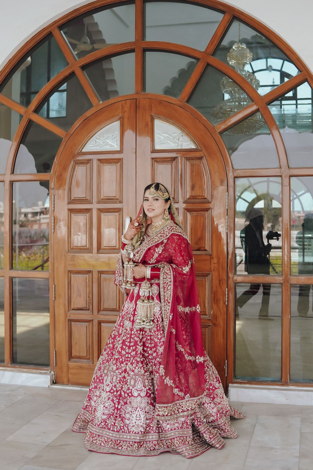 Indian bride in crimson red bridal lehenga with gold embroidery posing before grand wooden arched doors.