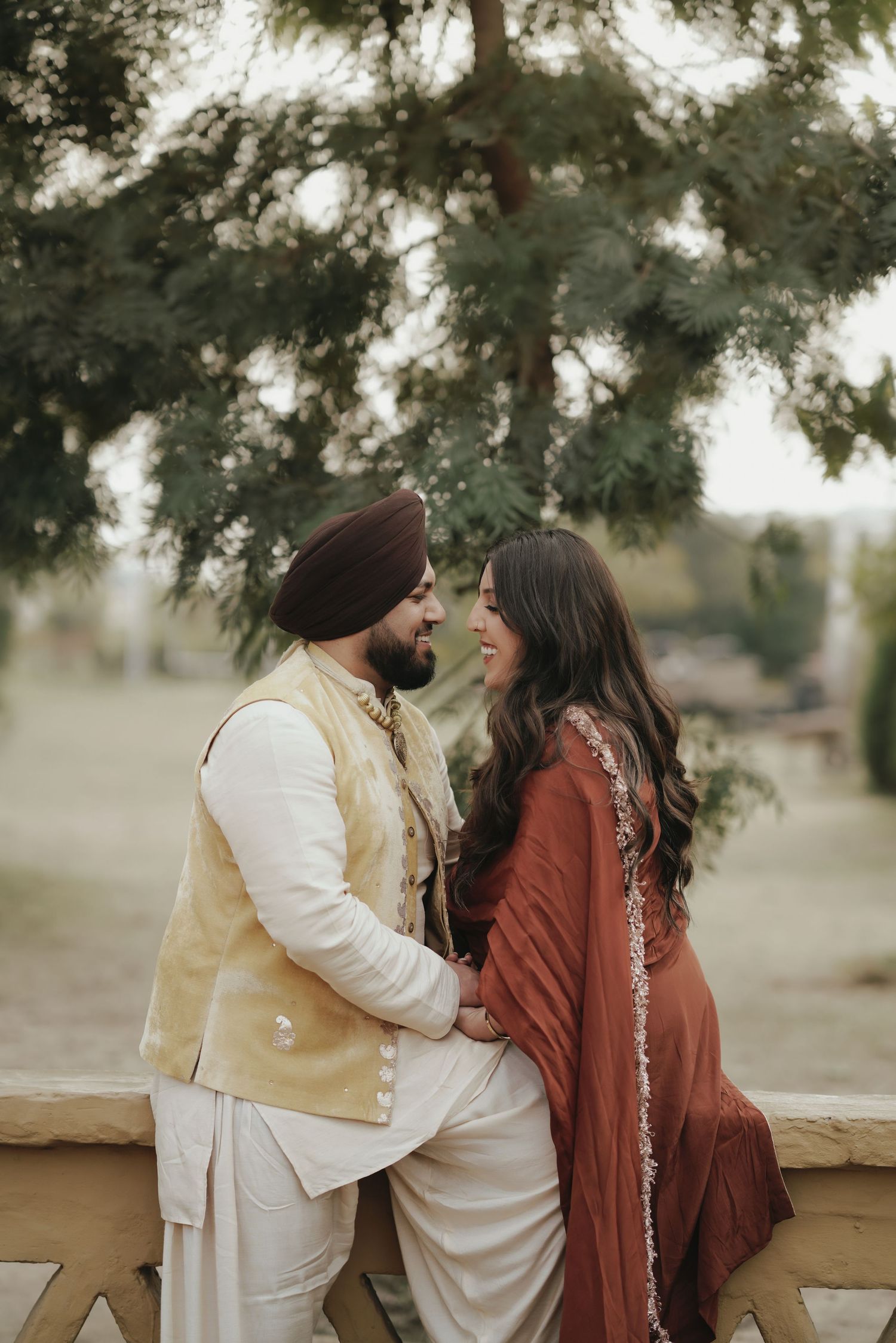 Sikh groom in turban and golden vest with bride in rust dupatta sharing a romantic moment outdoors.
