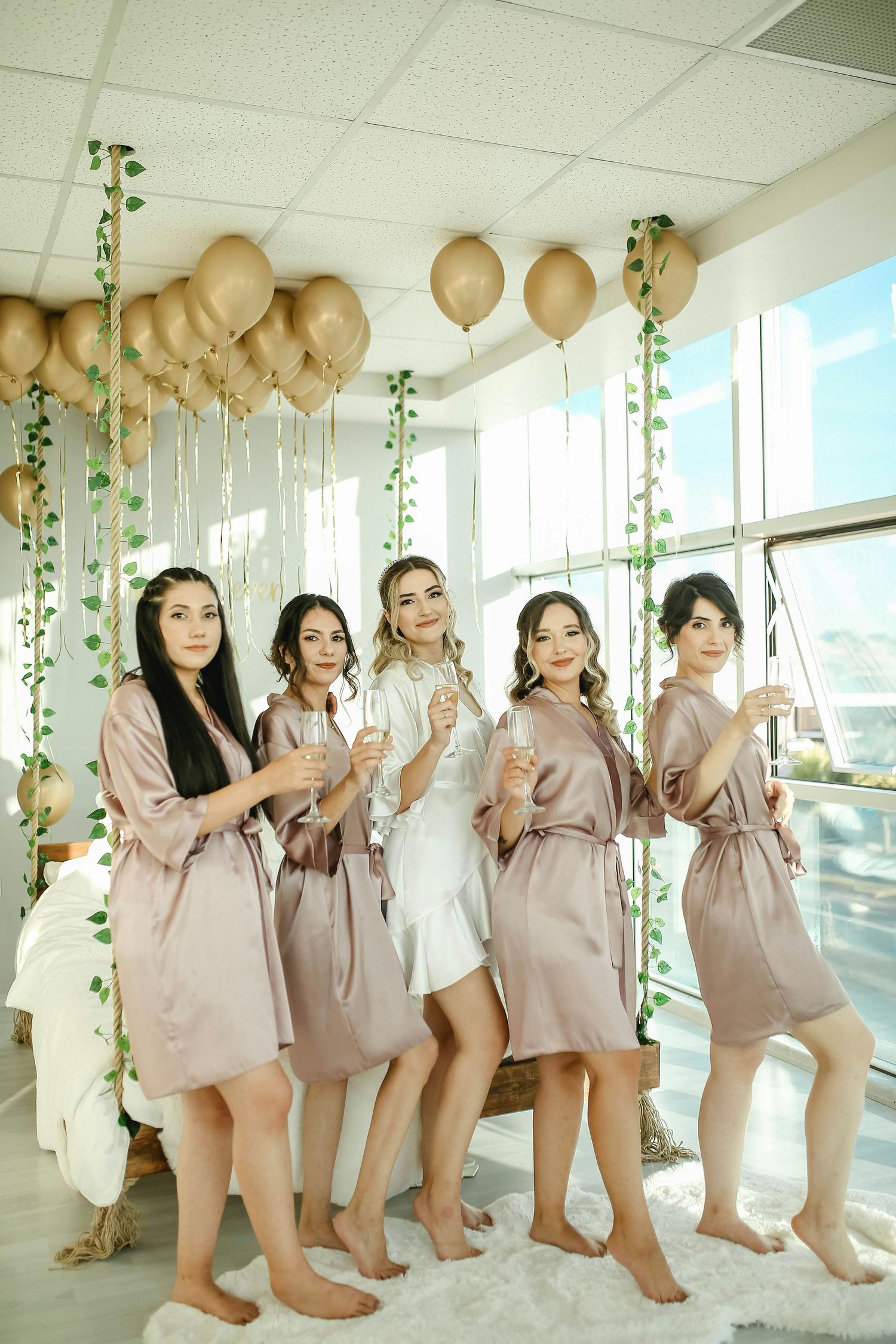 Bride and four bridesmaids in matching satin robes celebrating with champagne glasses at bridal party.