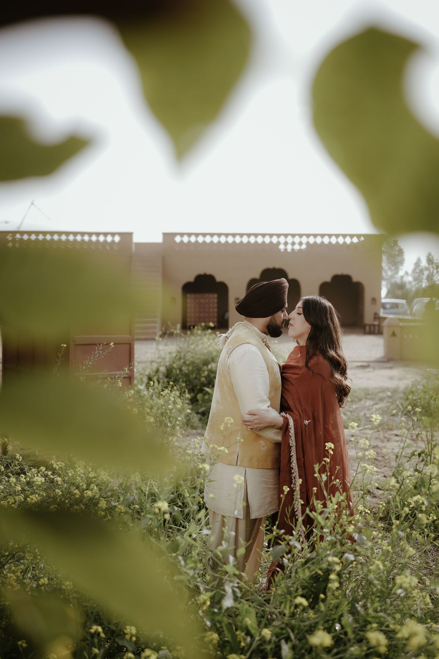 Sikh couple sharing a kiss in mustard flower field with Rajasthani architecture backdrop.