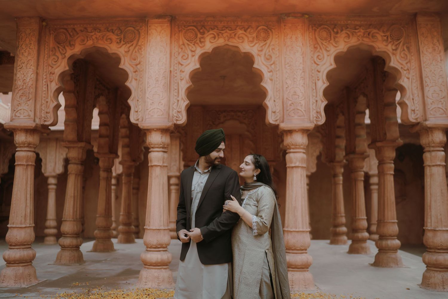Sikh couple in traditional attire sharing a romantic moment among ornate pink sandstone temple columns.