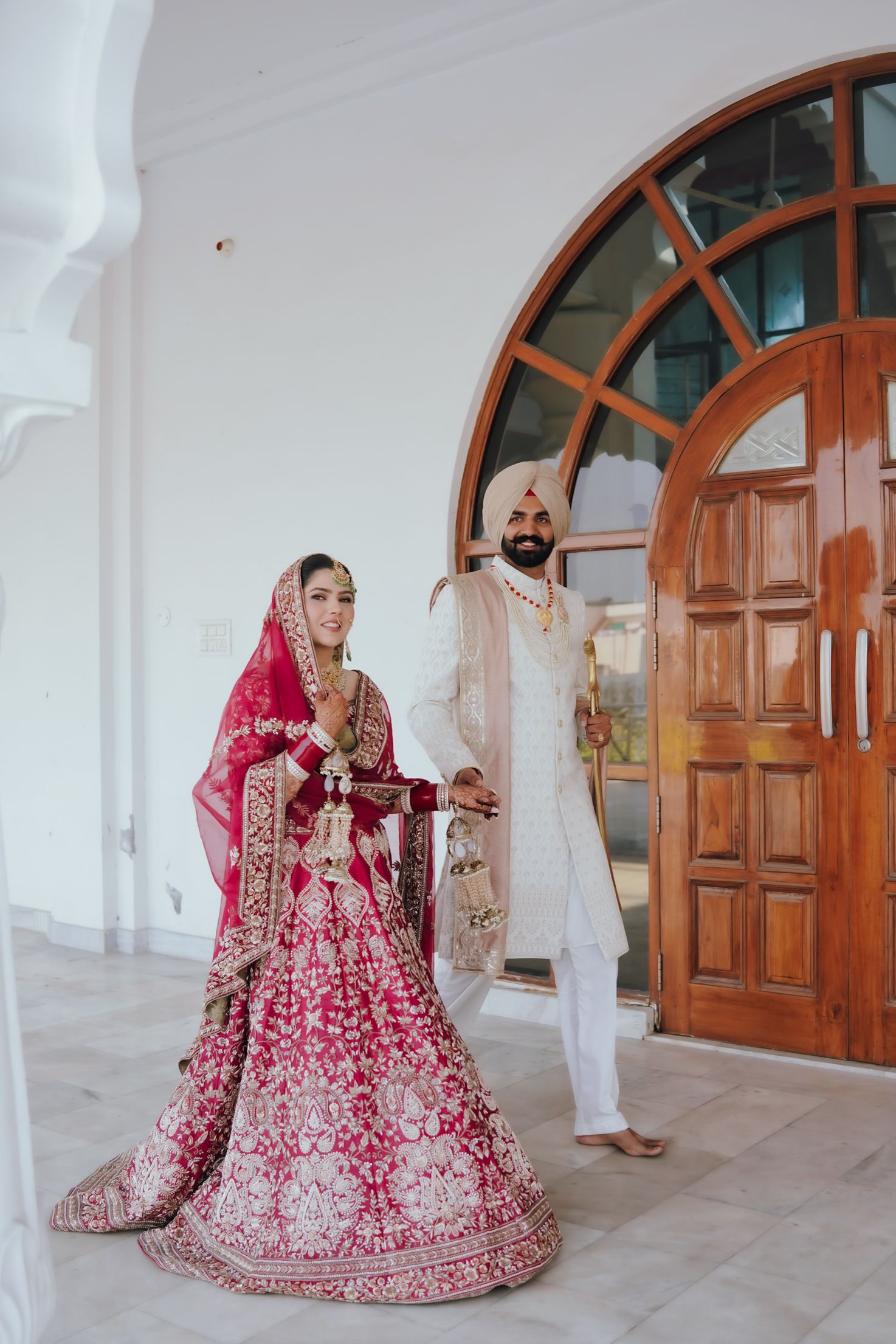 Indian Sikh bride in red lehenga and groom in white sherwani posing at ornate wooden doors.