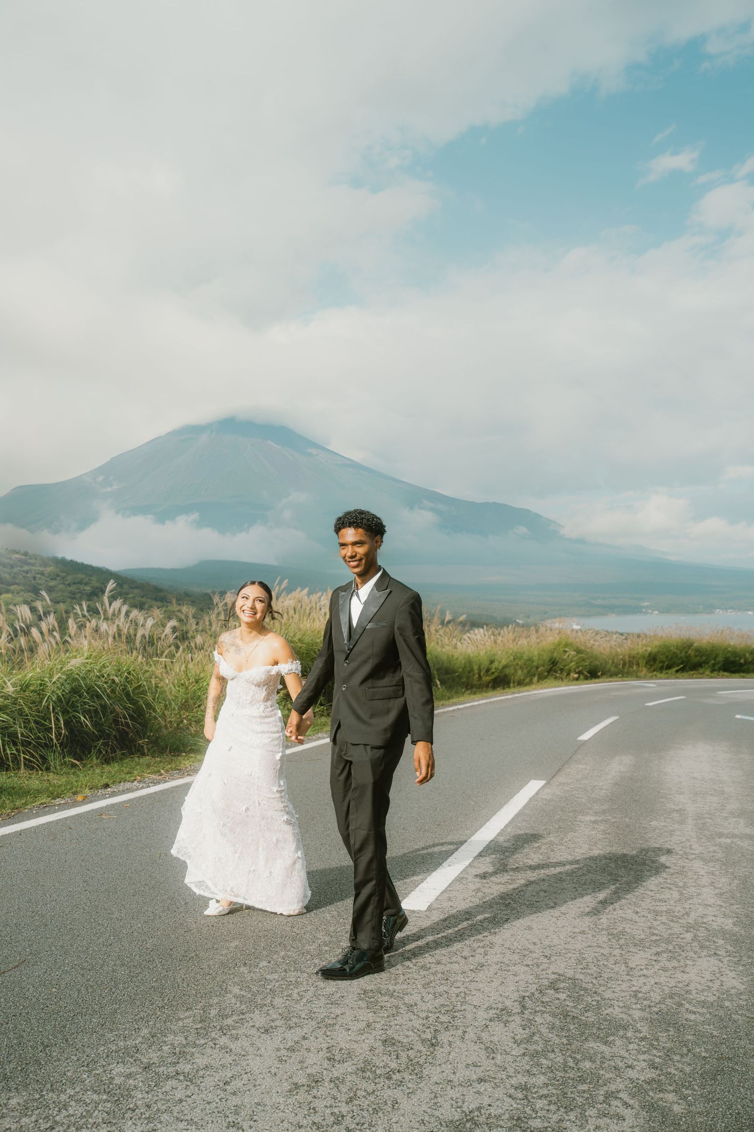 Interracial couple in wedding attire holding hands on a scenic road with a misty mountain backdrop.