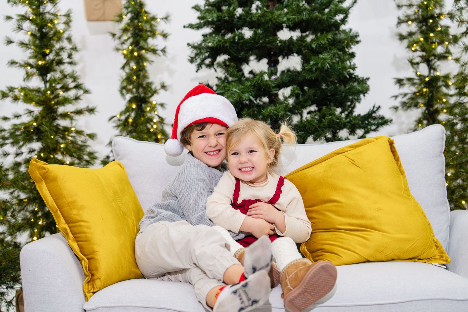 Two children in Santa hats hugging on a white sofa with yellow pillows and Christmas trees in the background.