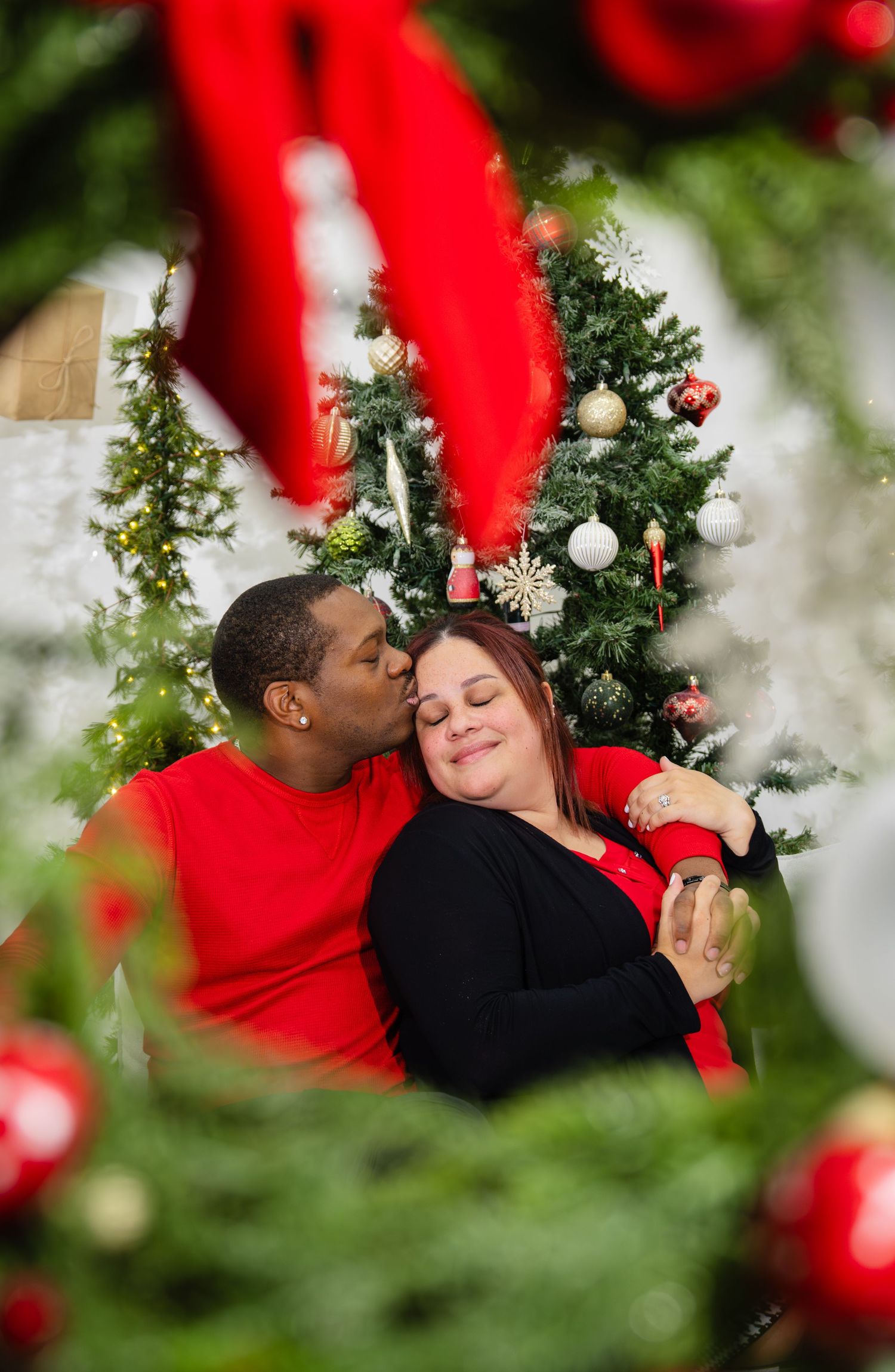 Romantic interracial couple sharing a kiss framed by red Christmas ornaments and a festive tree.