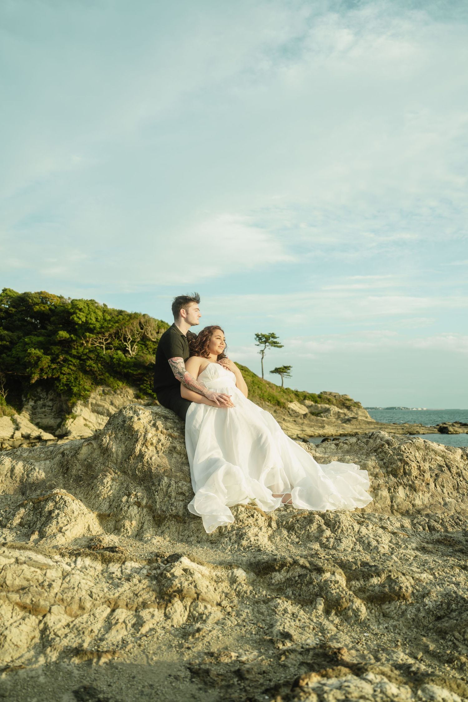 Couple in wedding attire sitting closely on rocky coastal cliffs with a serene ocean view behind them.