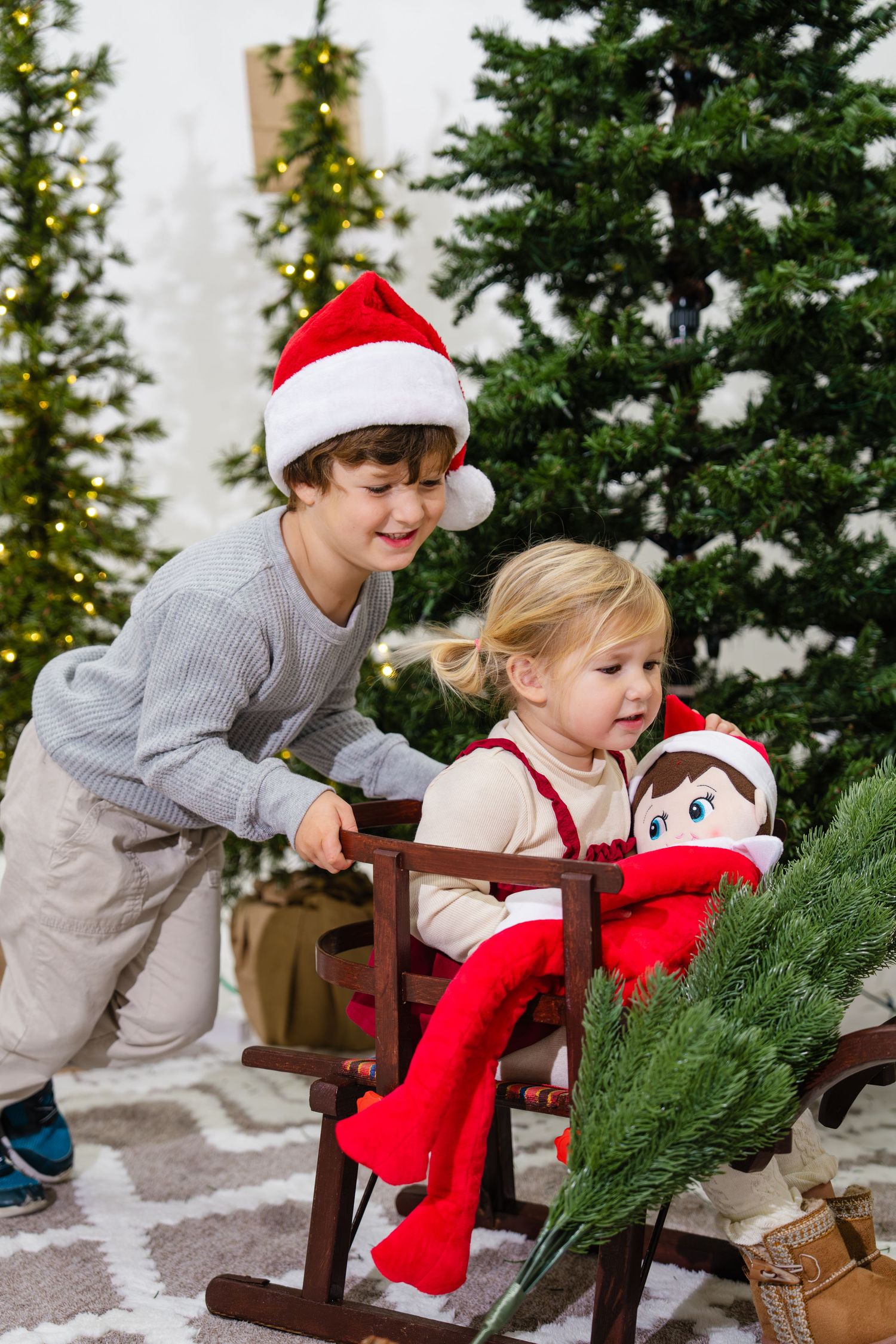 Boy in Santa hat pushing a little girl on a wooden sled near decorated Christmas trees.