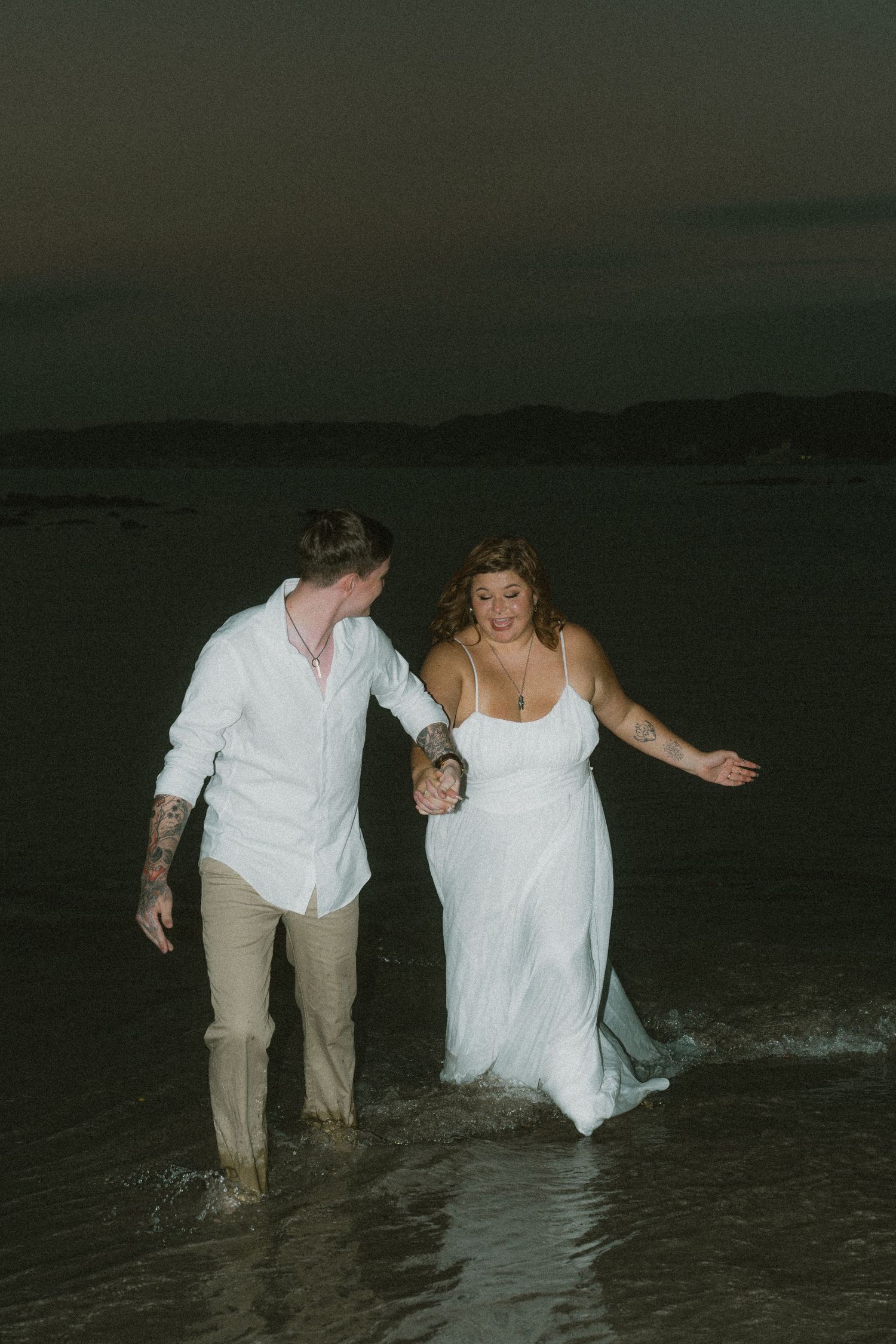 Couple in wedding attire laughing and wading through ocean waves together at night on the beach.