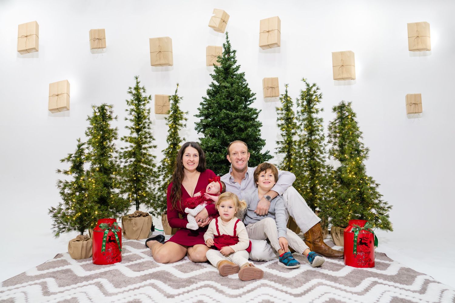 Happy family of five posing in front of Christmas trees with gift boxes on a white wall backdrop.