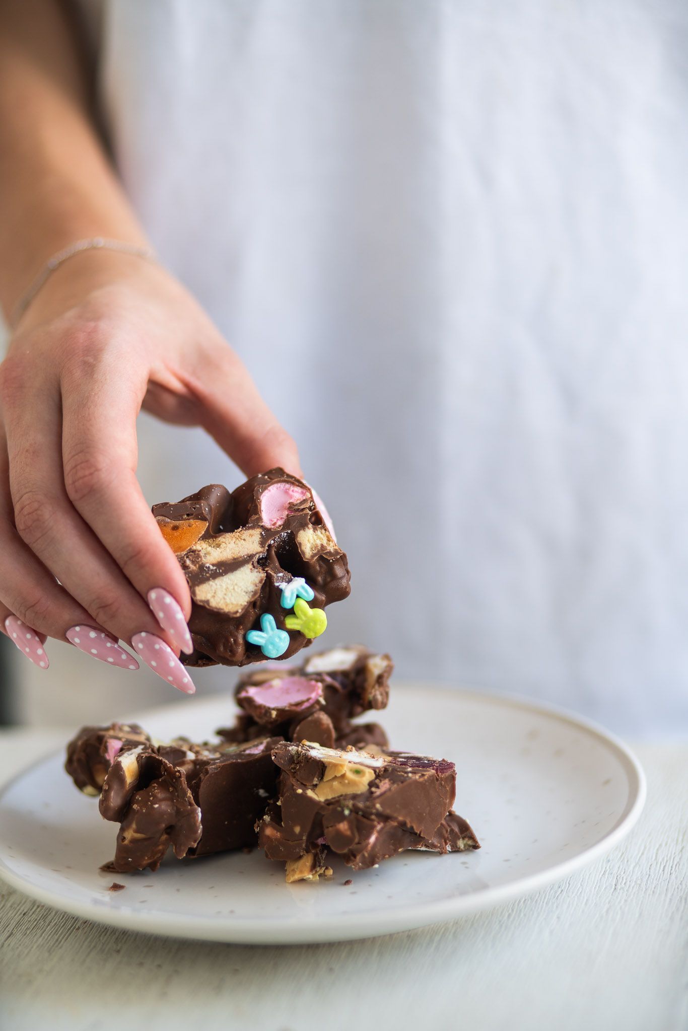 A woman's hand with pink nails picks up a piece of chocolate rocky road bark topped with colorful Easter bunny sprinkles from a white plate.