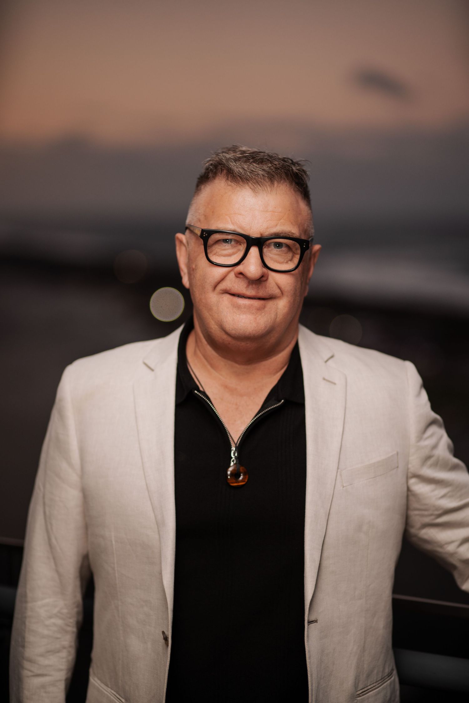 Middle-aged man in white blazer and black shirt wearing bold glasses and pendant necklace, smiling at sunset.