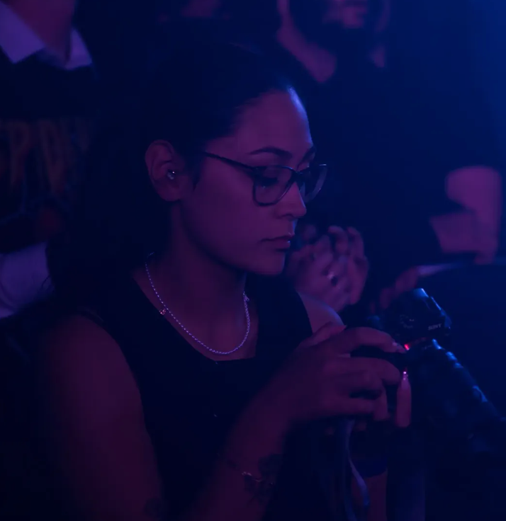 Woman with glasses holding a microphone in a dimly lit venue with purple lighting.