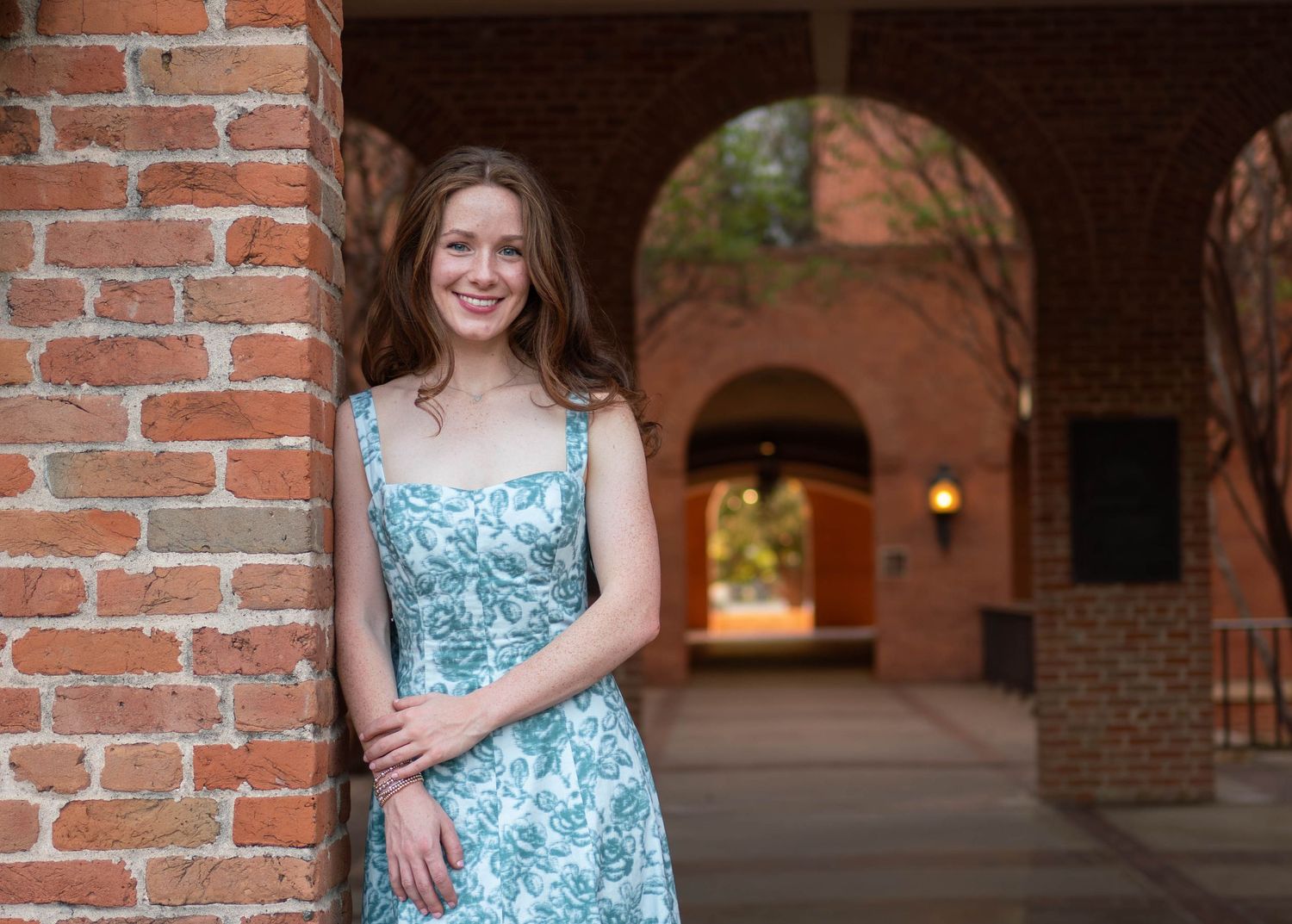 Smiling young woman in floral dress leaning against brick wall under arched university corridor.