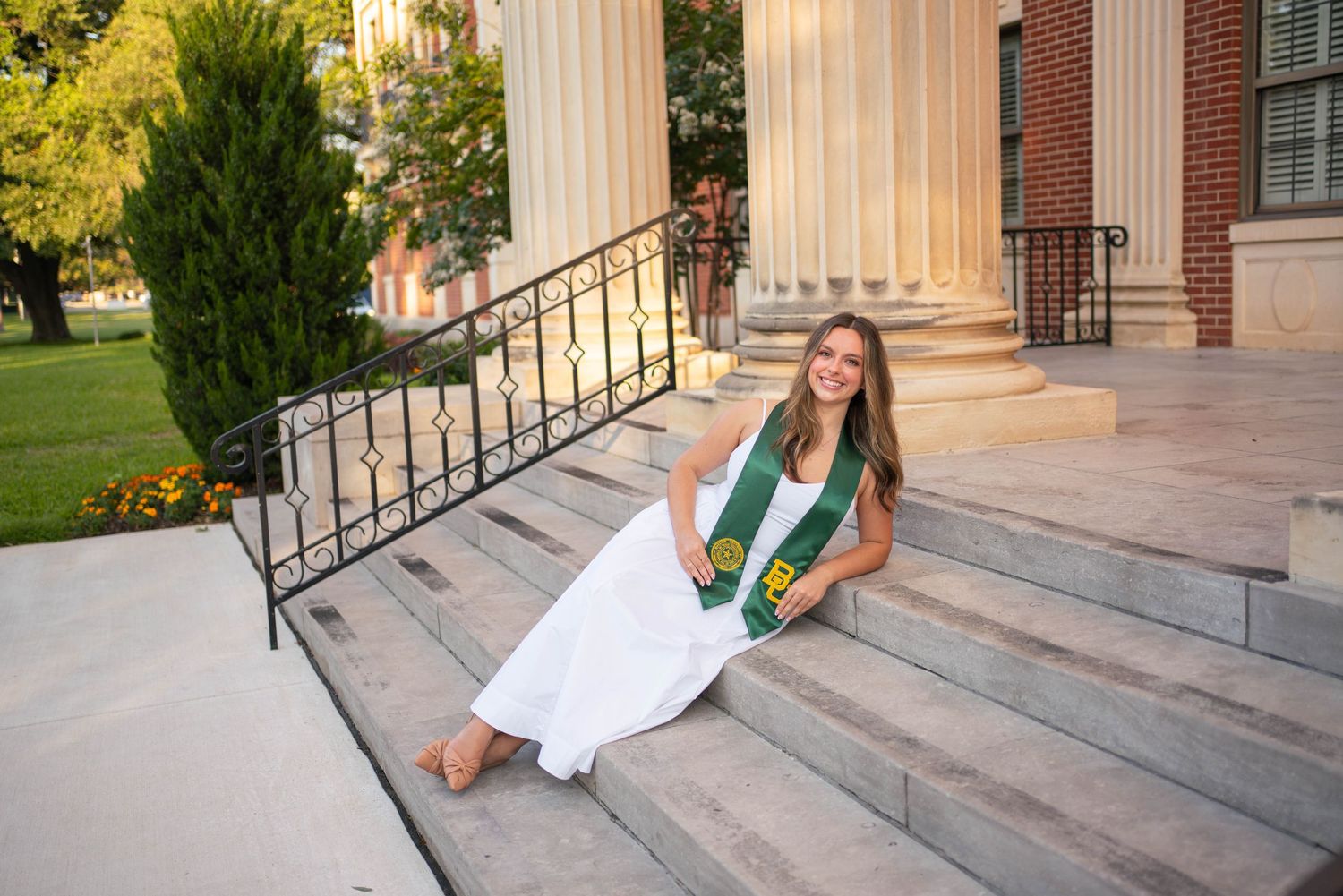 Young woman in white dress and green graduation stole sitting on university building steps for grad photos.