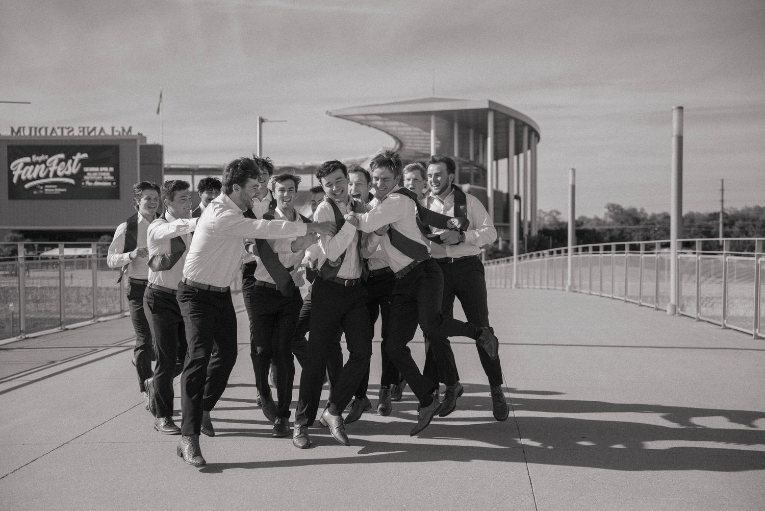 Black and white photo of a lively group of groomsmen walking and joking together on a bridge.