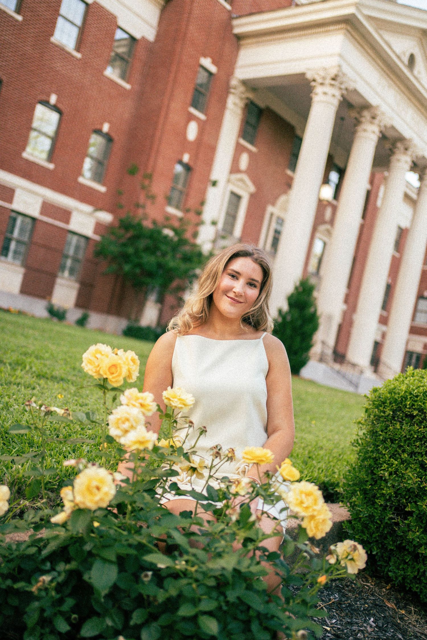 Young woman in white dress smiling among yellow roses outside brick building with white columns.