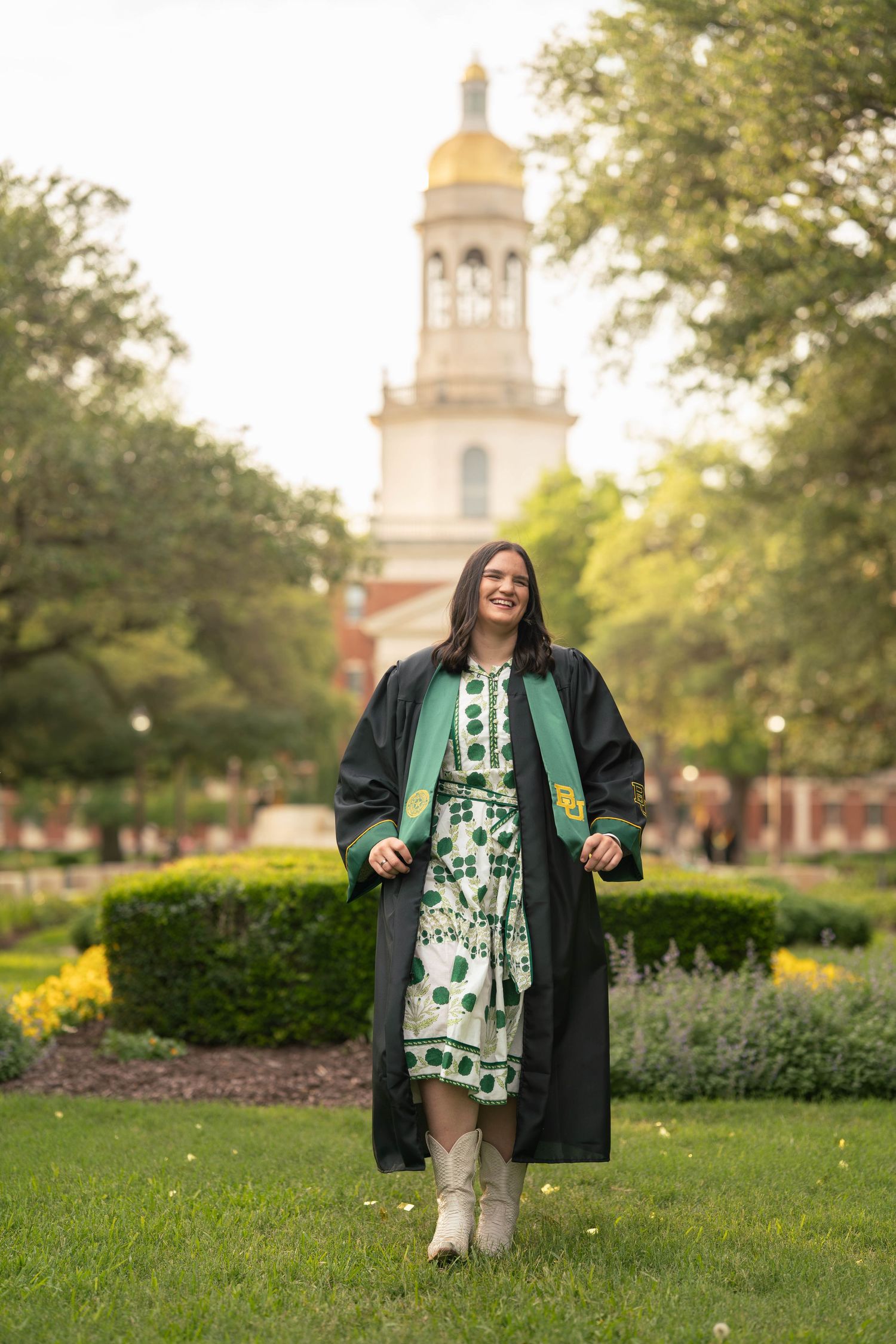 A smiling graduate in black regalia and floral dress walks on green campus lawn with a gold-domed bell tower behind her.