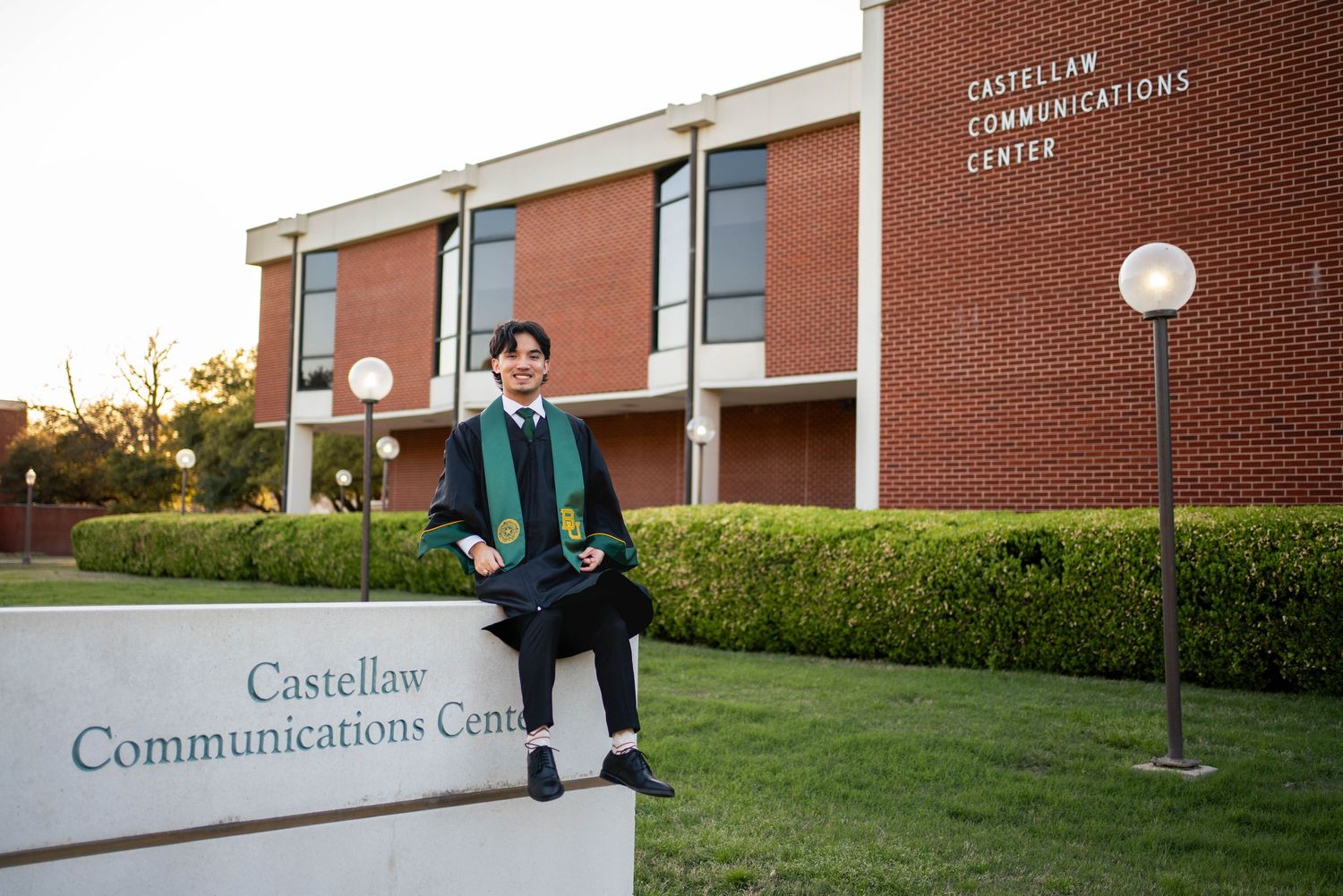 Baylor University graduate in green gown sitting on Castellaw Communications Center sign at dusk.