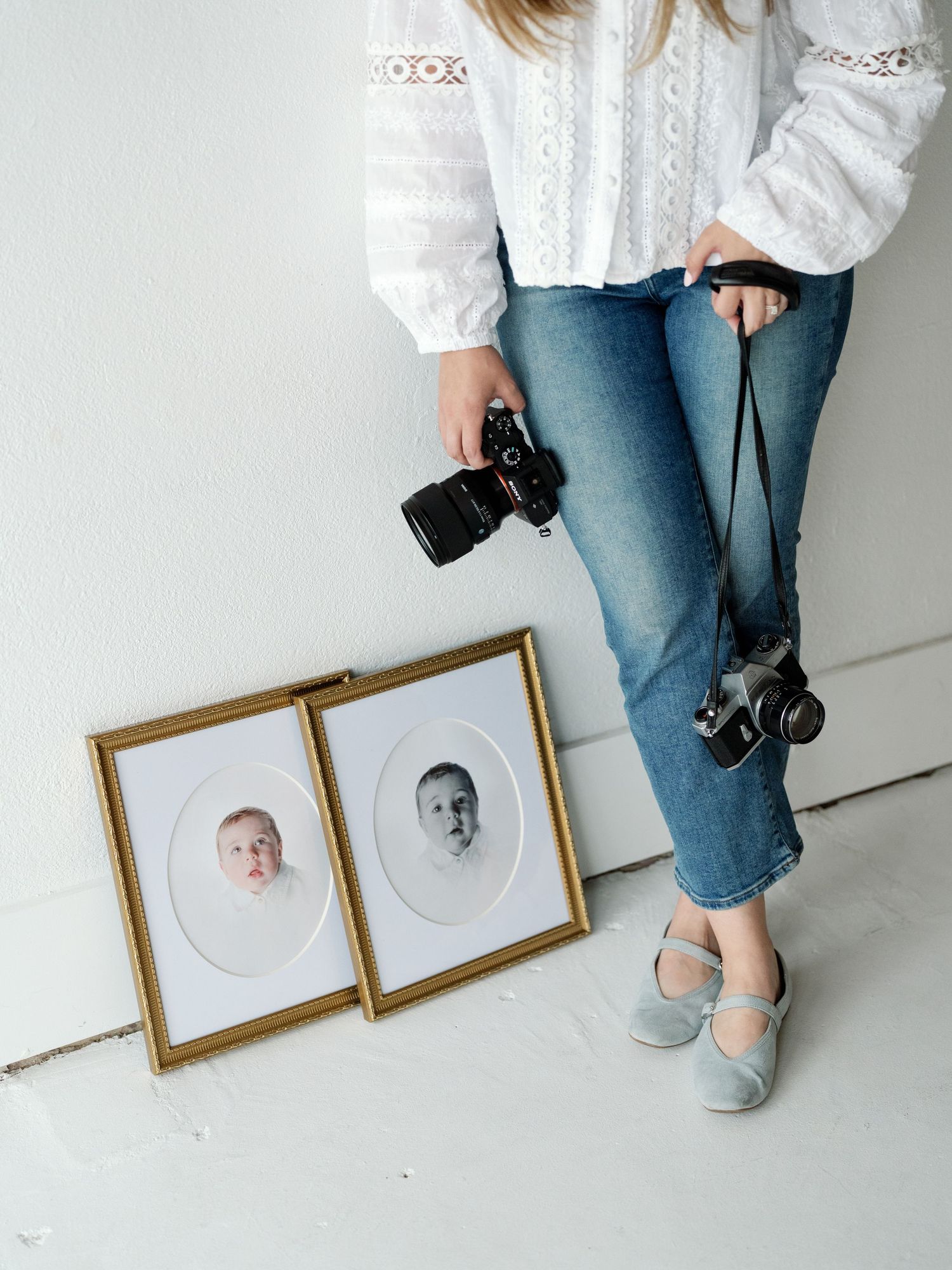 Photographer holding two cameras beside framed baby portrait prints leaning against a white wall, wearing jeans and a white blouse.