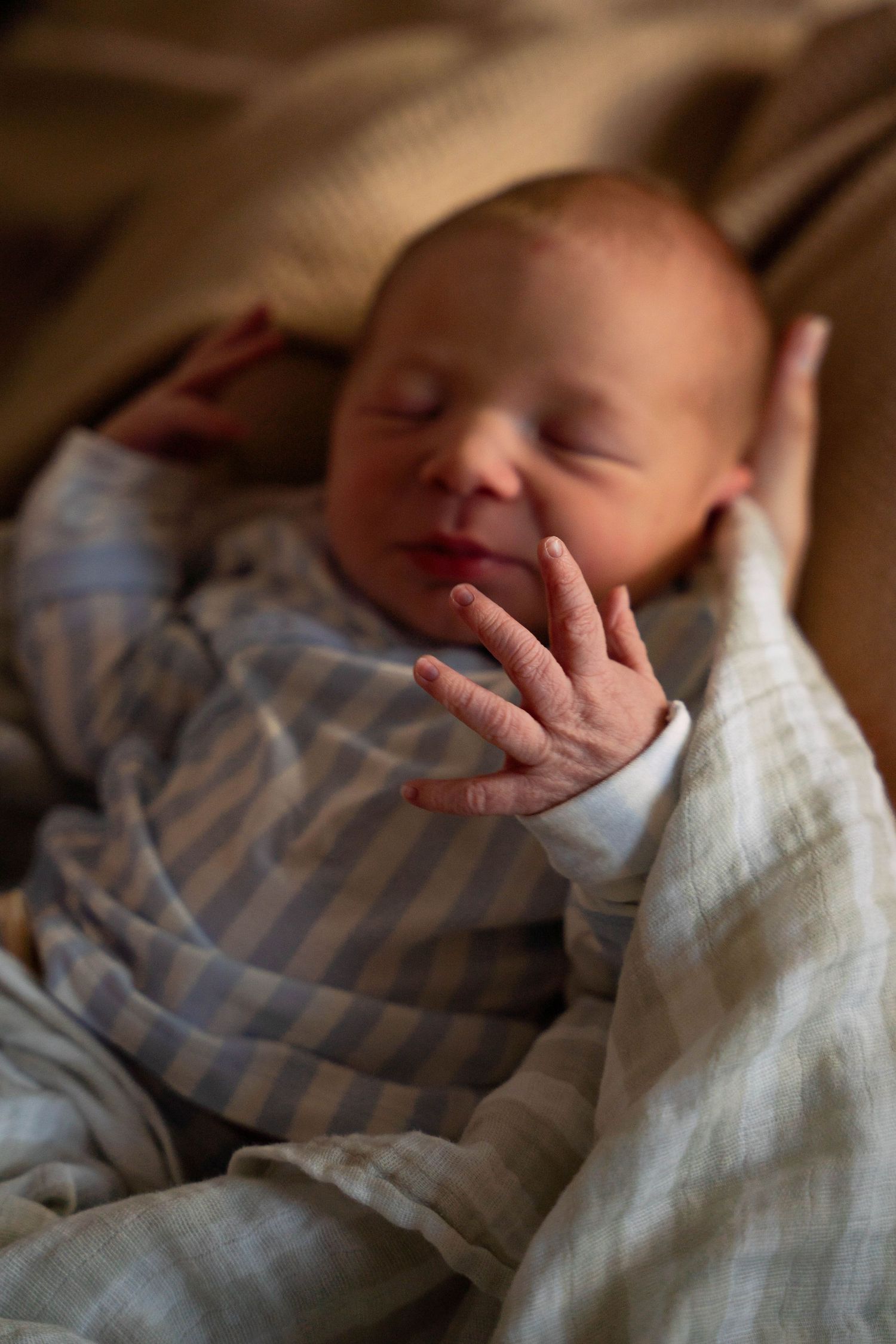Sleeping newborn baby in striped onesie with tiny fingers raised, wrapped in soft muslin blanket.