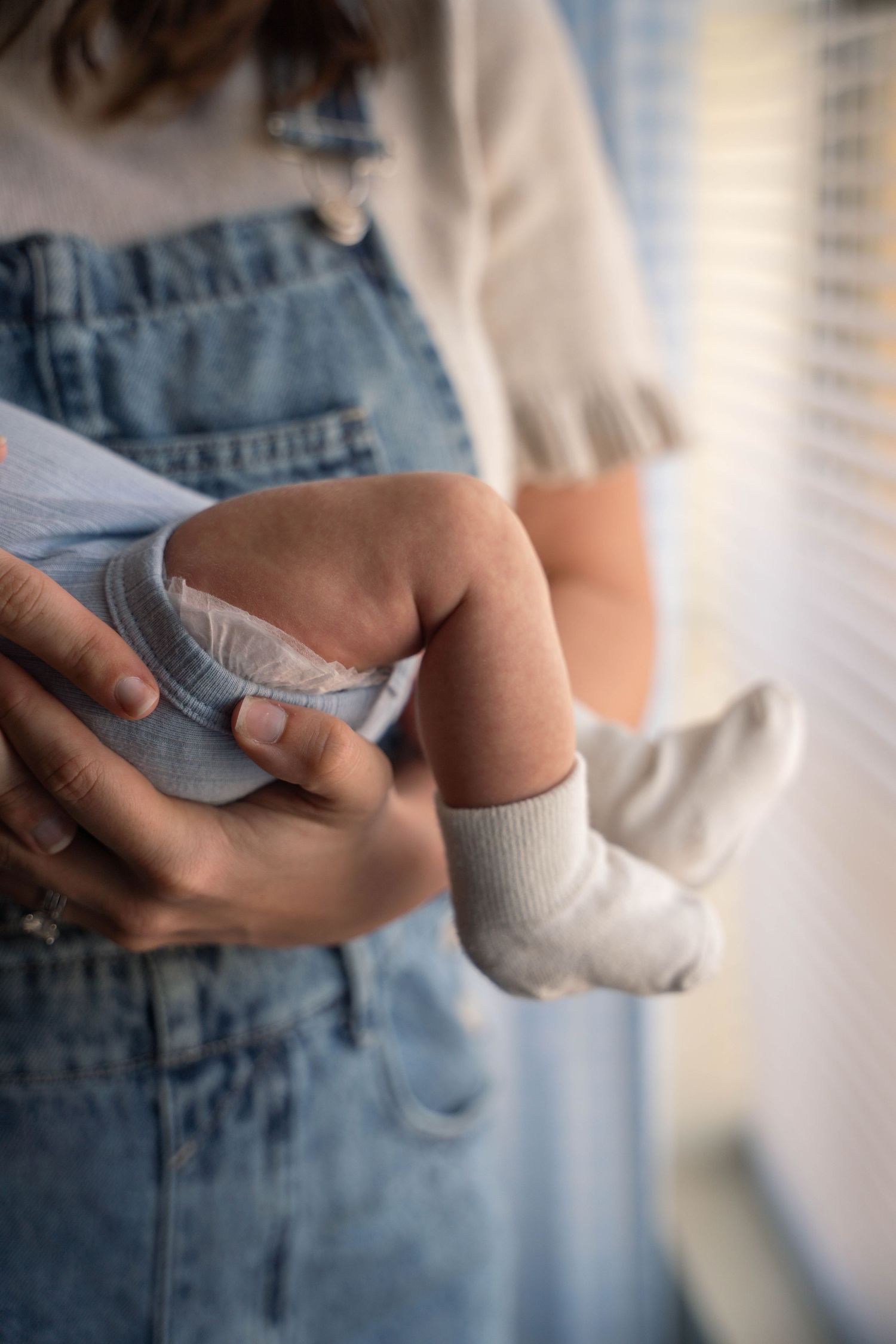 Close-up of mother in denim overalls holding newborn baby legs and feet near window.