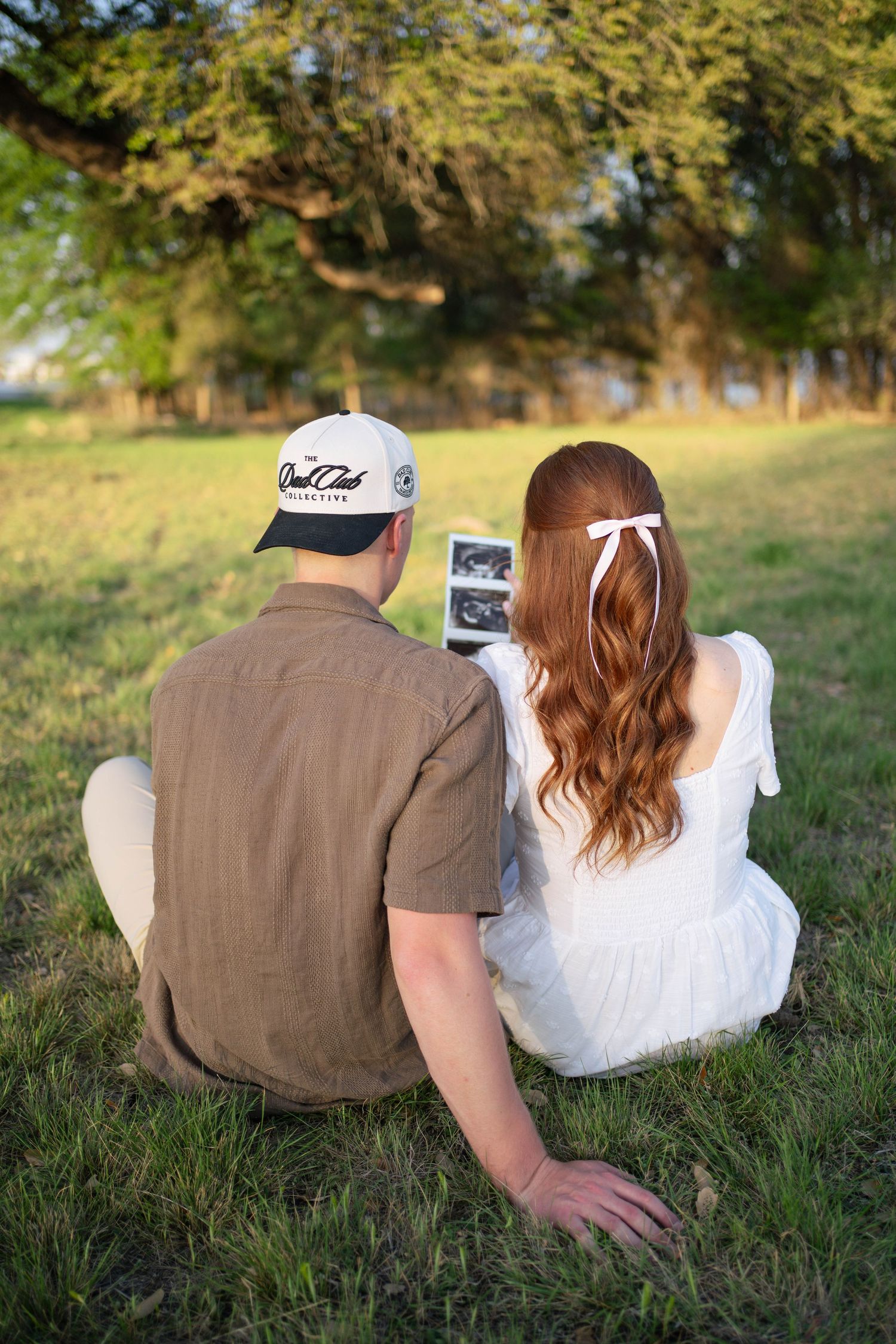 Couple sitting on grass viewing ultrasound photo, woman in white dress with bow, man in brown shirt and cap.