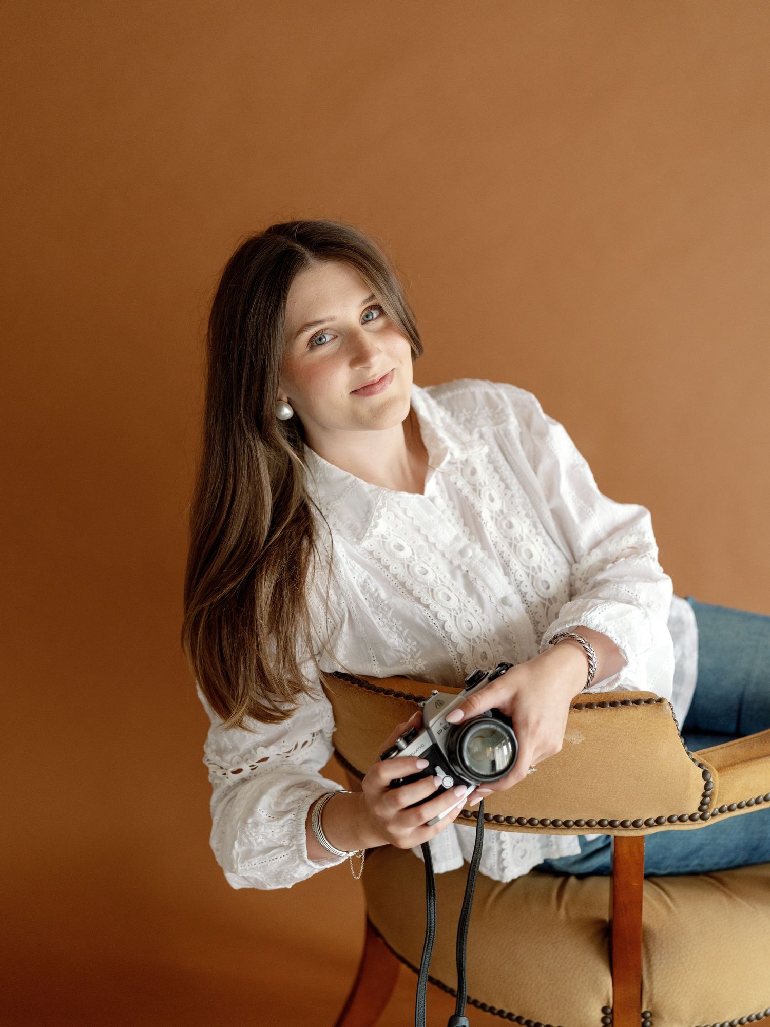 Woman with long brown hair holds a camera while sitting on a gold chair against a warm brown background.