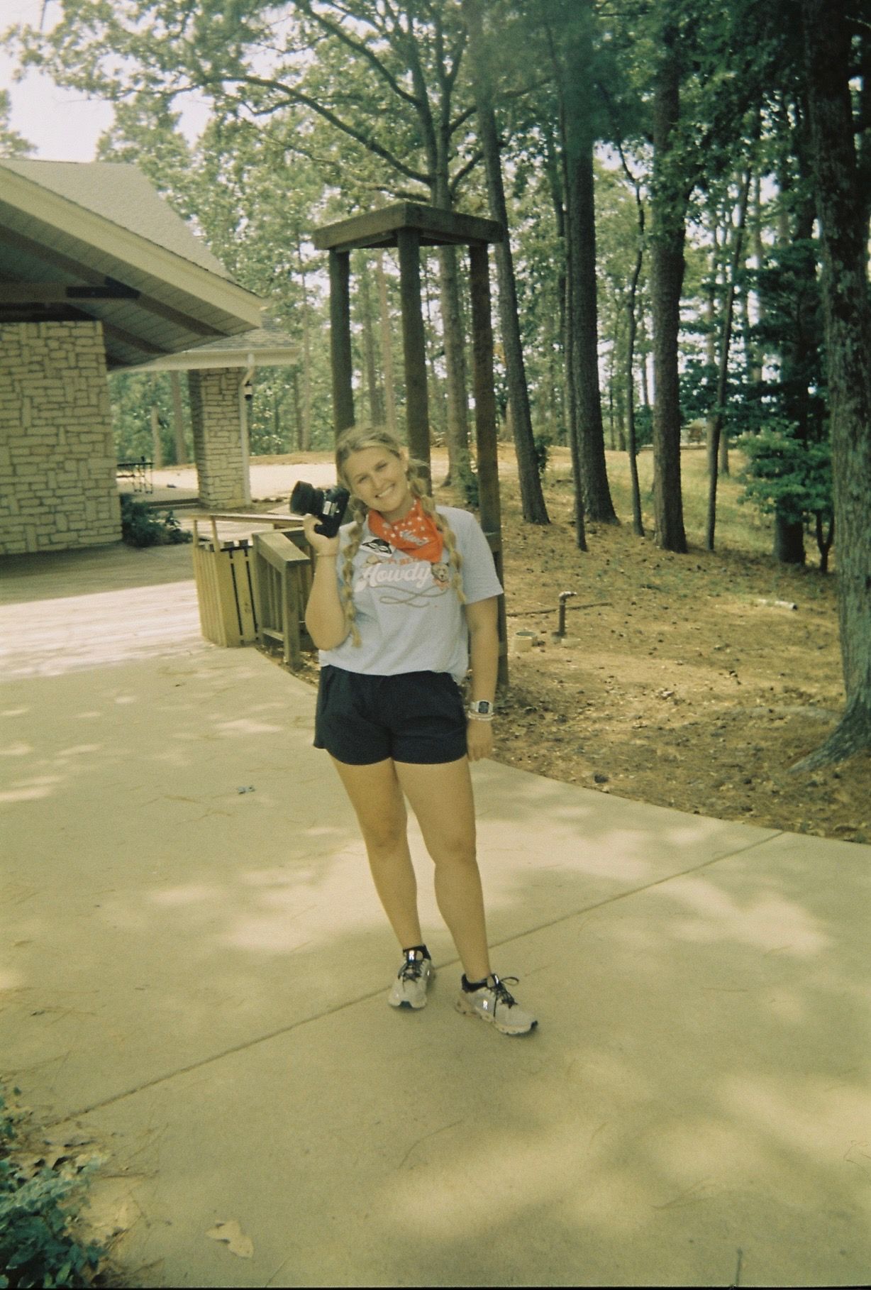 Young woman smiling and holding a camera wearing shorts and a bandana outdoors near brick building.