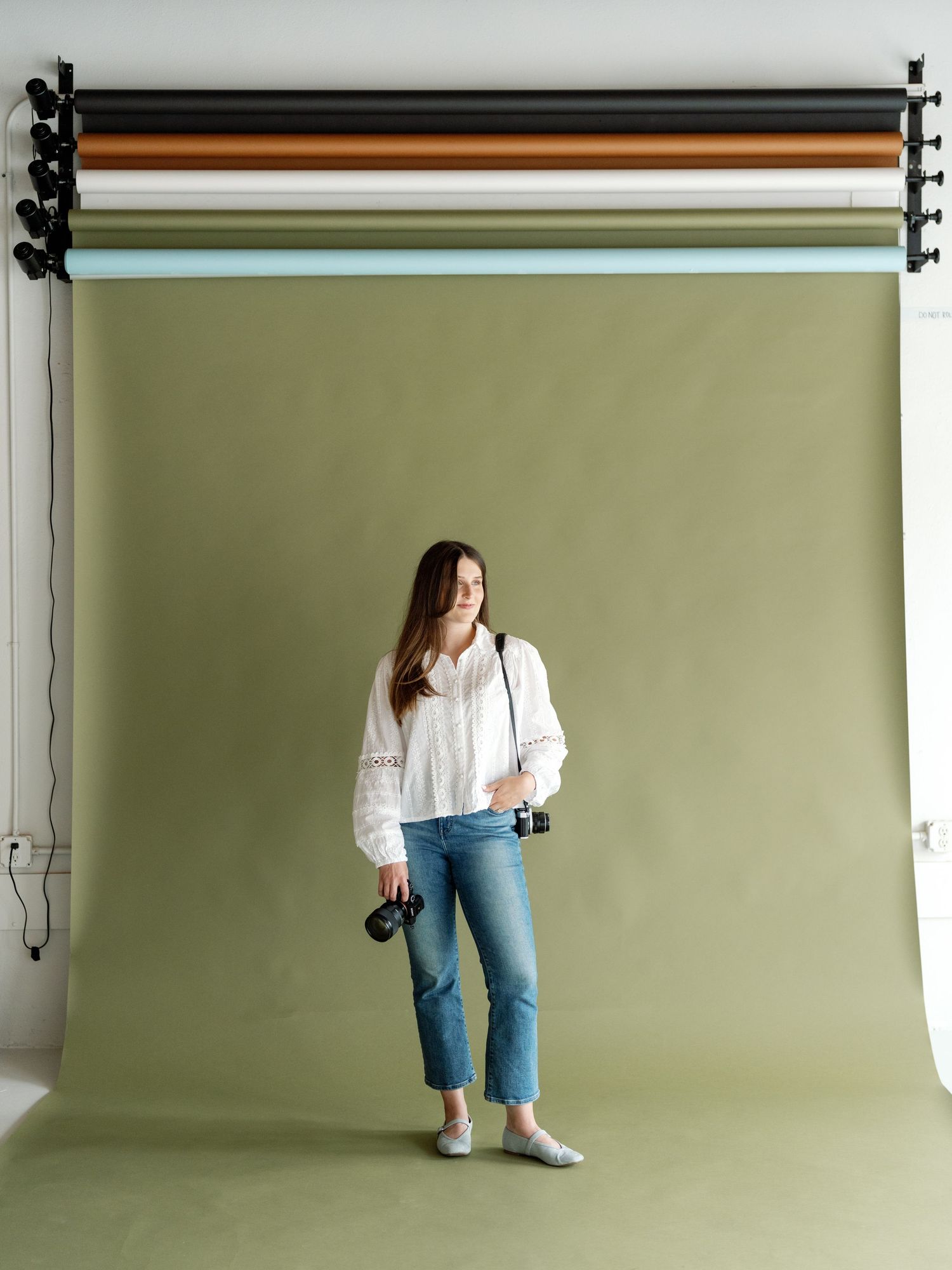 Photographer standing in a studio holding cameras, wearing white blouse and jeans, in front of a sage green backdrop.