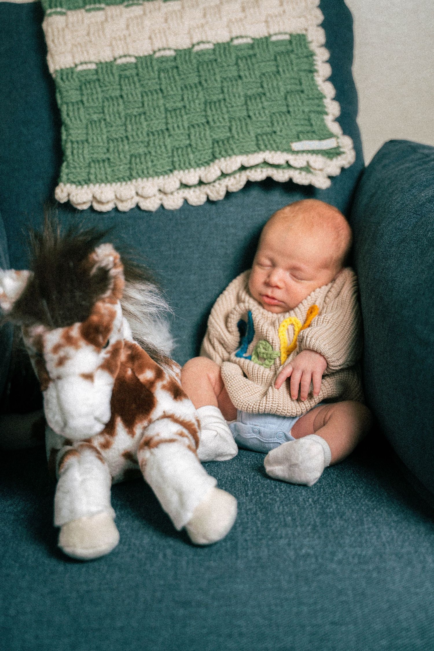 Sleeping newborn baby in knit sweater sits on teal sofa beside stuffed horse toy with crochet blanket nearby.