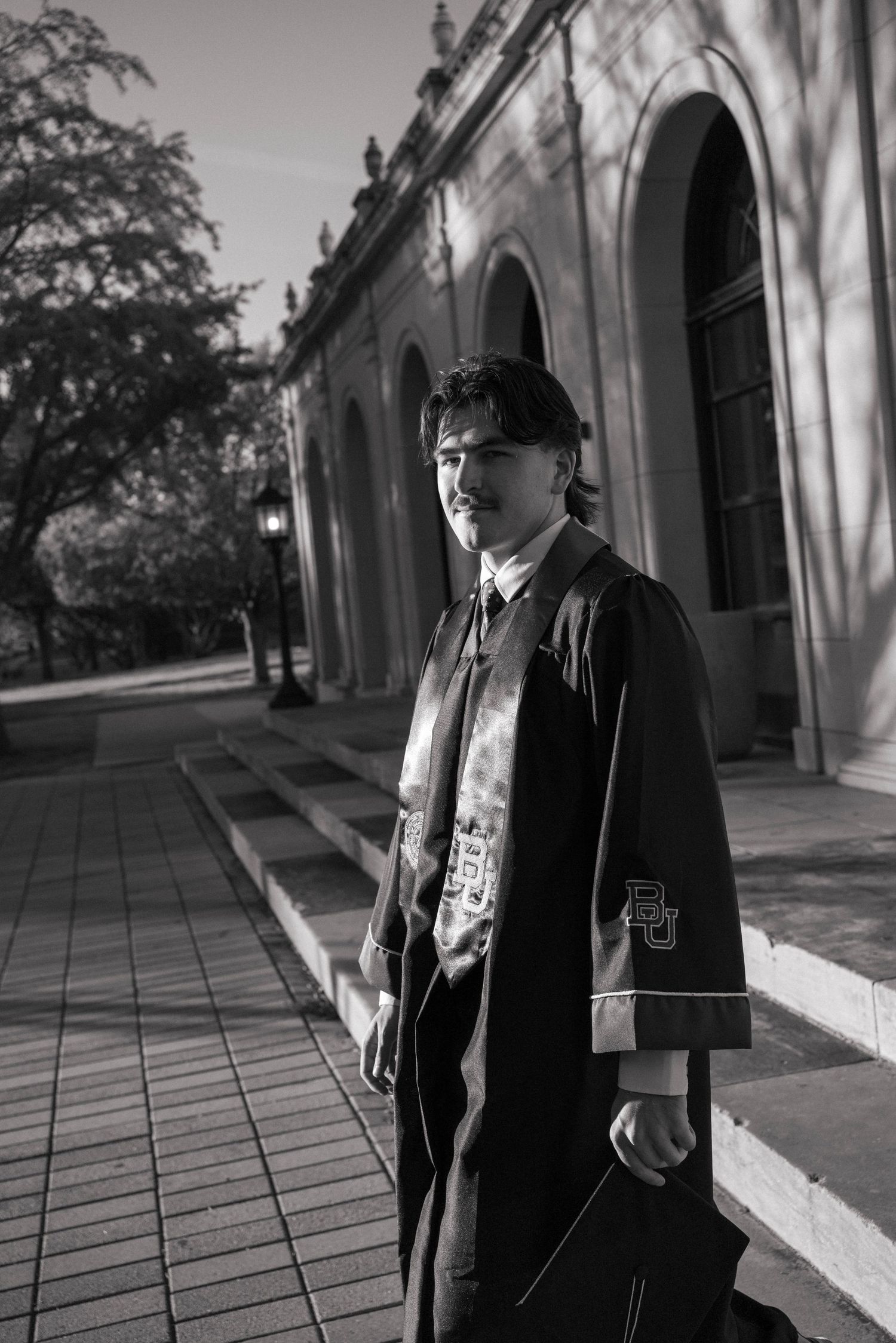 Black and white graduation portrait of a young man in cap and gown outside a historic building.