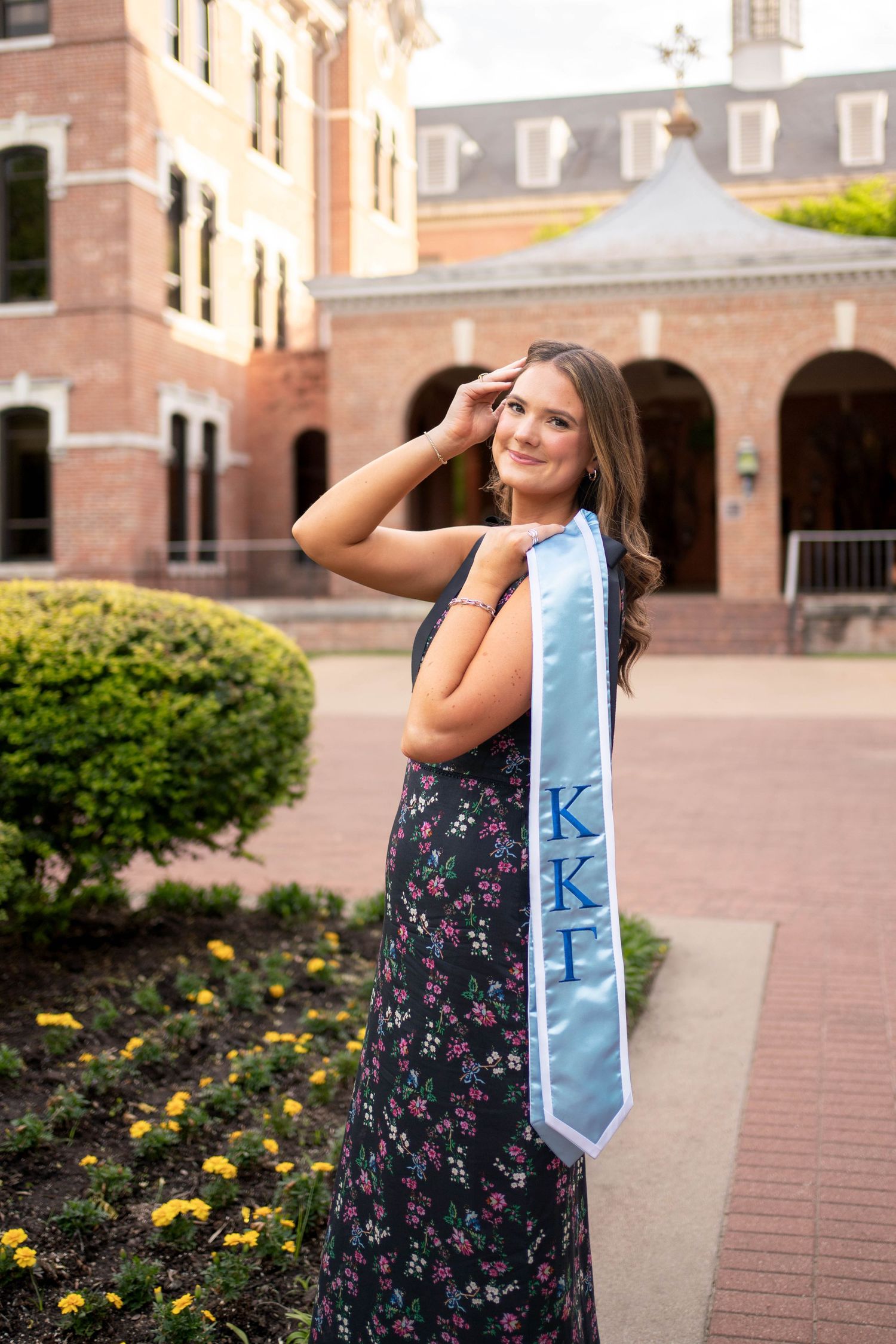Young woman in floral dress holding light blue Kappa Kappa Gamma graduation stole on campus.