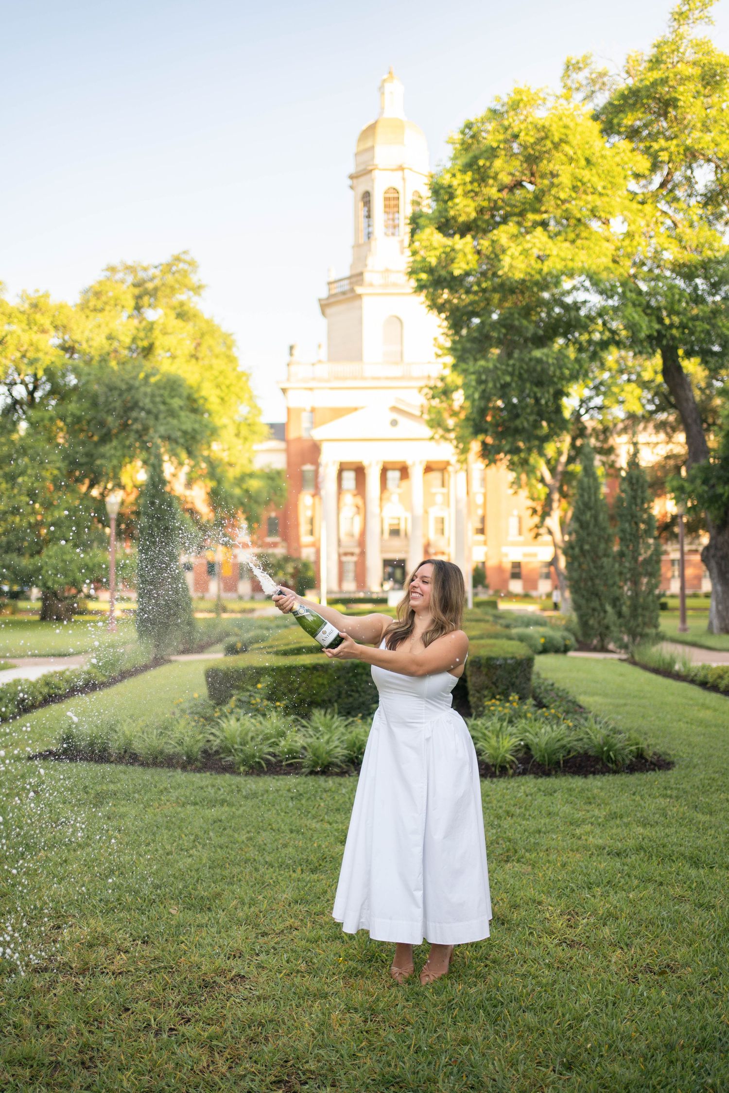 Woman in white dress popping champagne in front of Baylor University chapel at golden hour.