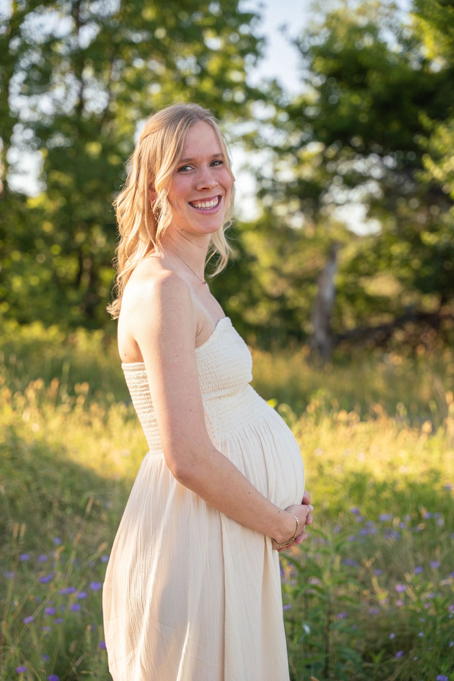 Smiling pregnant woman in cream dress cradling baby bump in a sunlit green field.