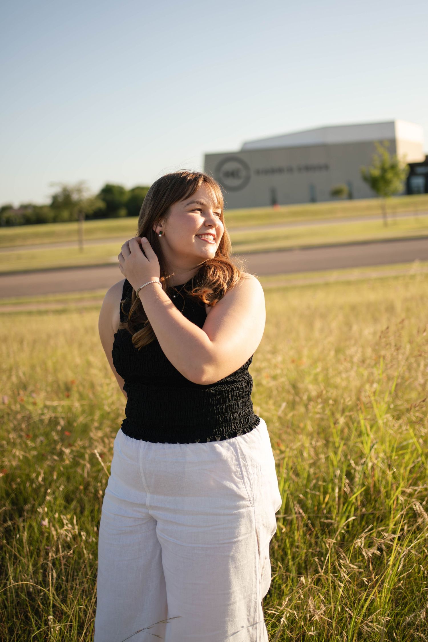 Young woman in black top and white pants smiling in a golden field at sunset.