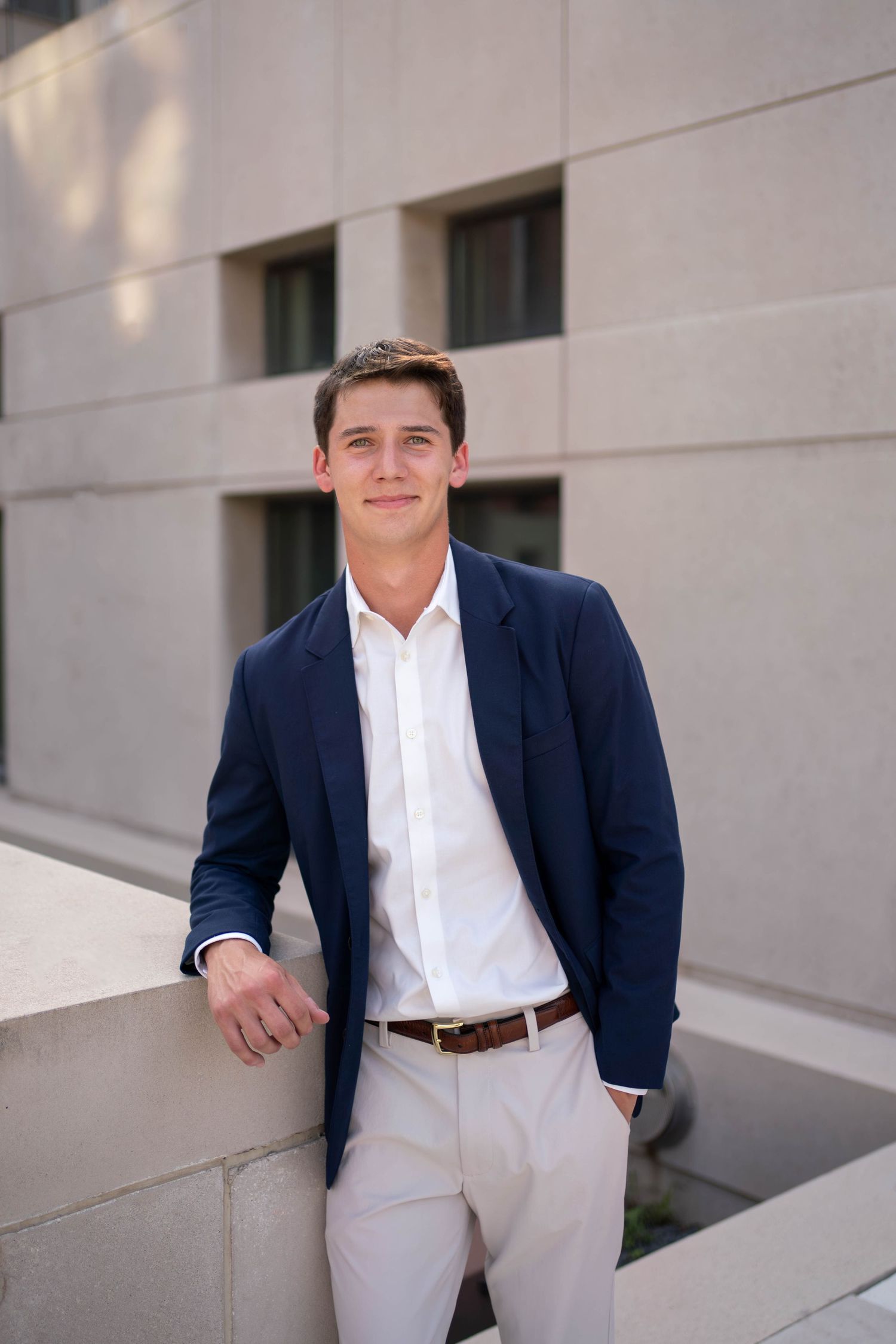 Young man in navy blazer and white shirt leaning against stone wall outside modern building.