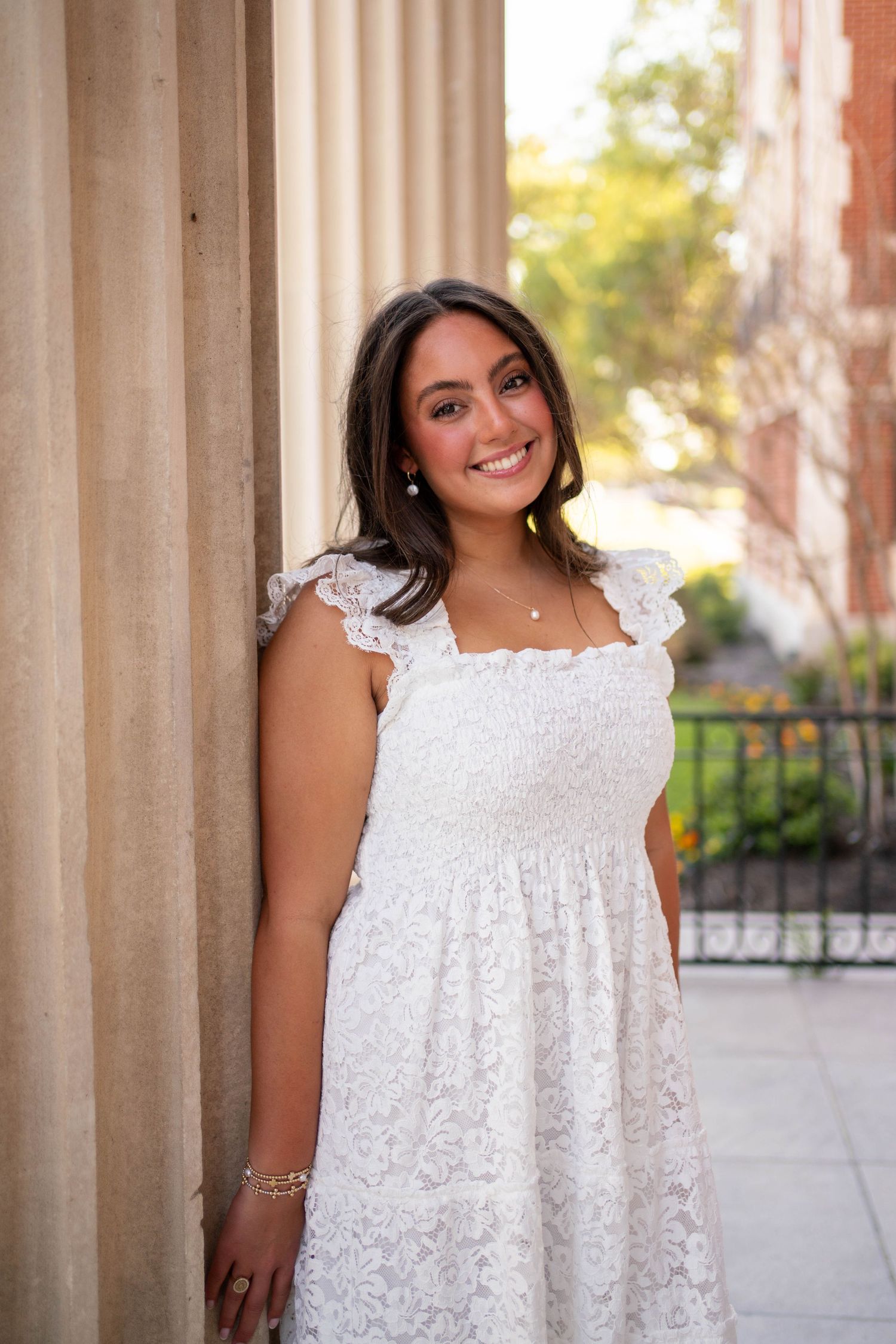 Young woman in white lace dress smiling beside stone column outdoors.