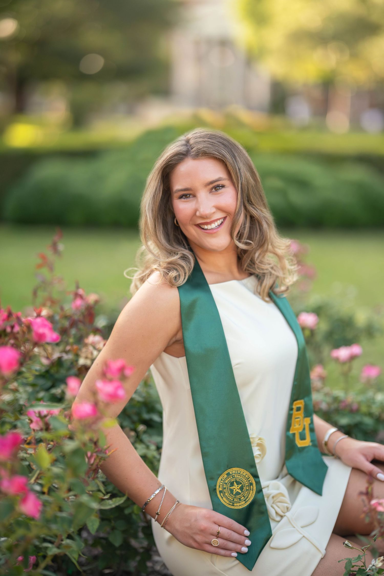 Smiling graduate in white dress wearing green and gold Baylor University graduation stole near pink roses.