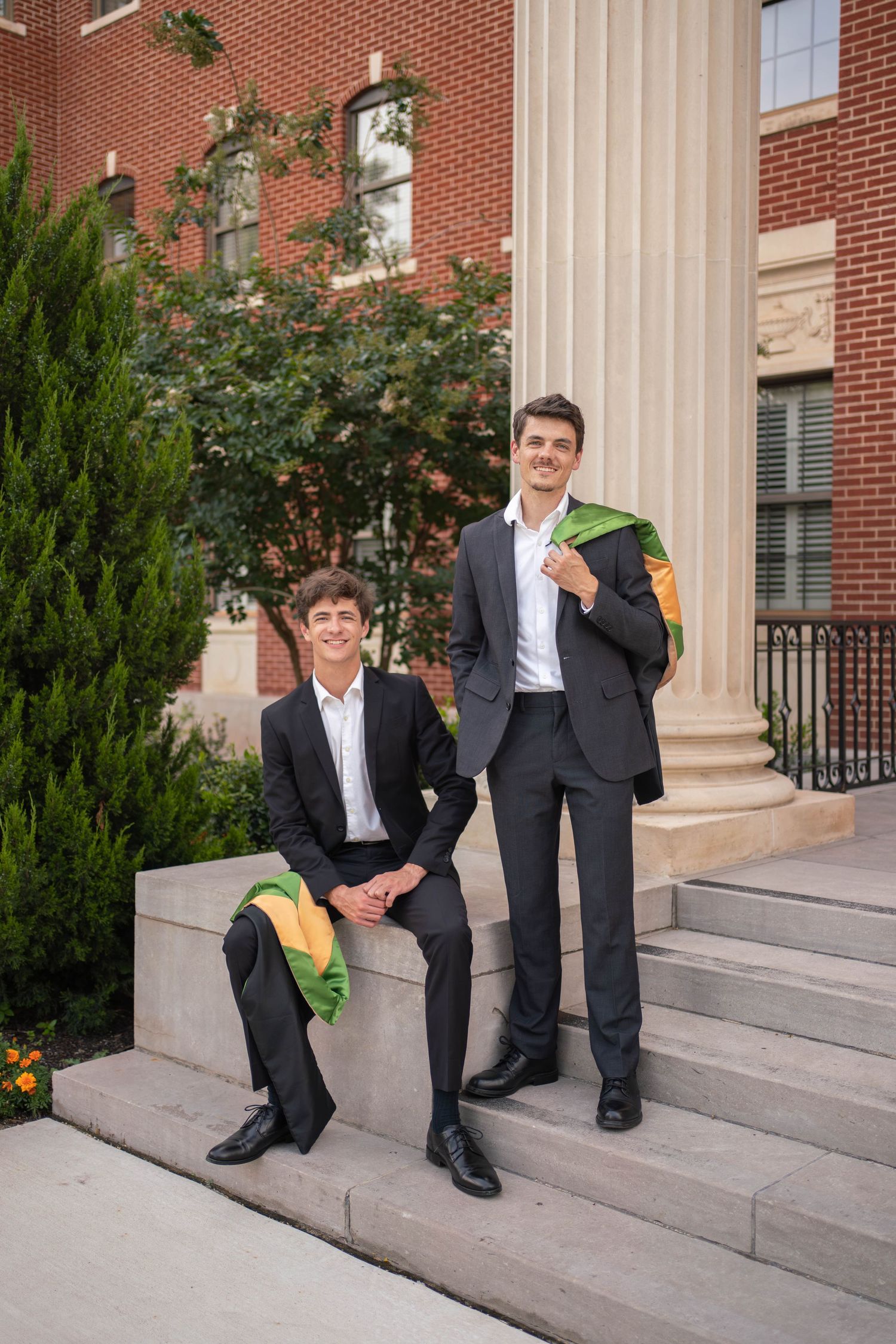 Two young men in black suits with graduation stoles posing on steps of brick building.