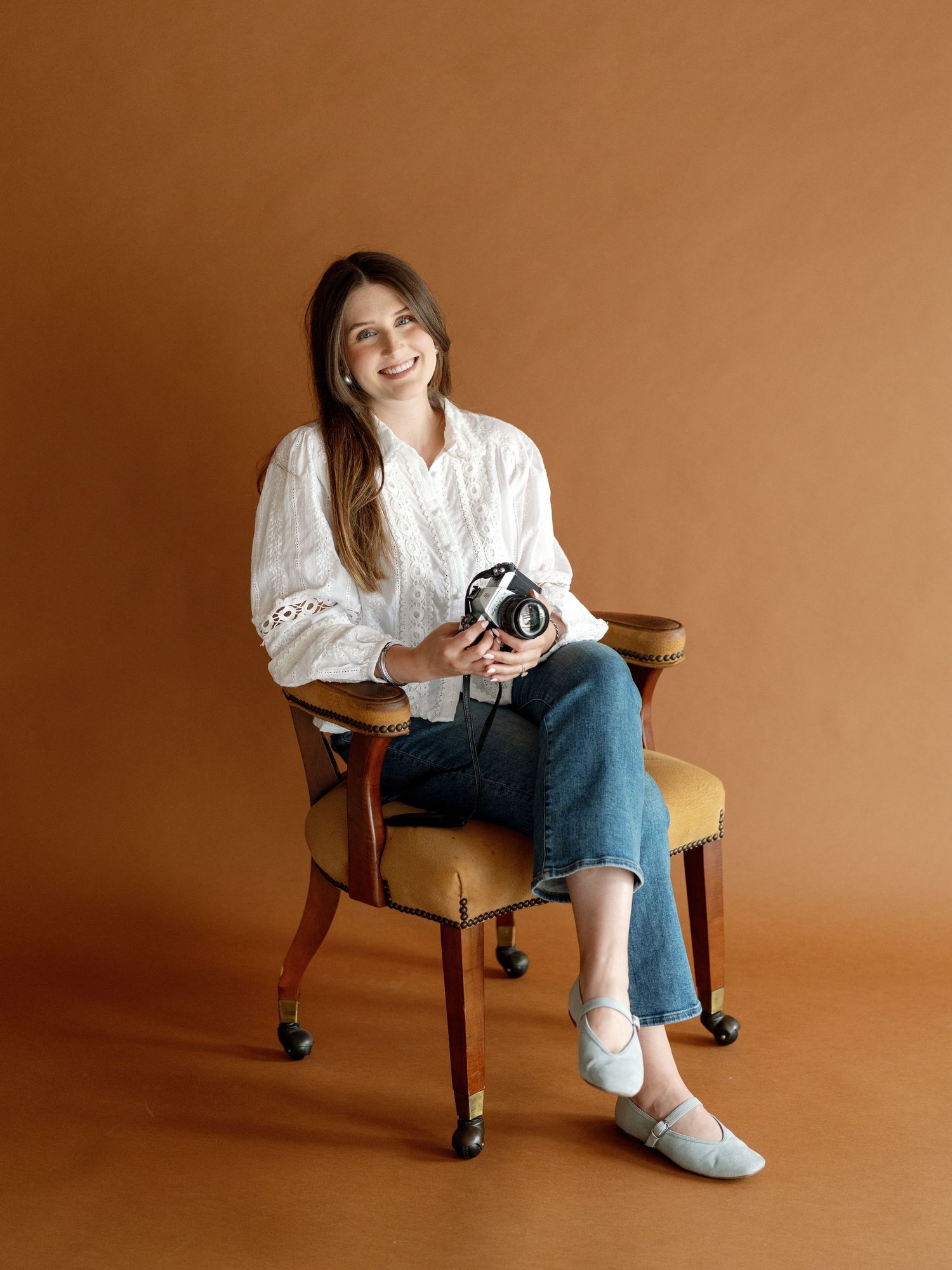 Smiling woman in white blouse and jeans holding a camera, seated on a vintage chair against a warm brown background.