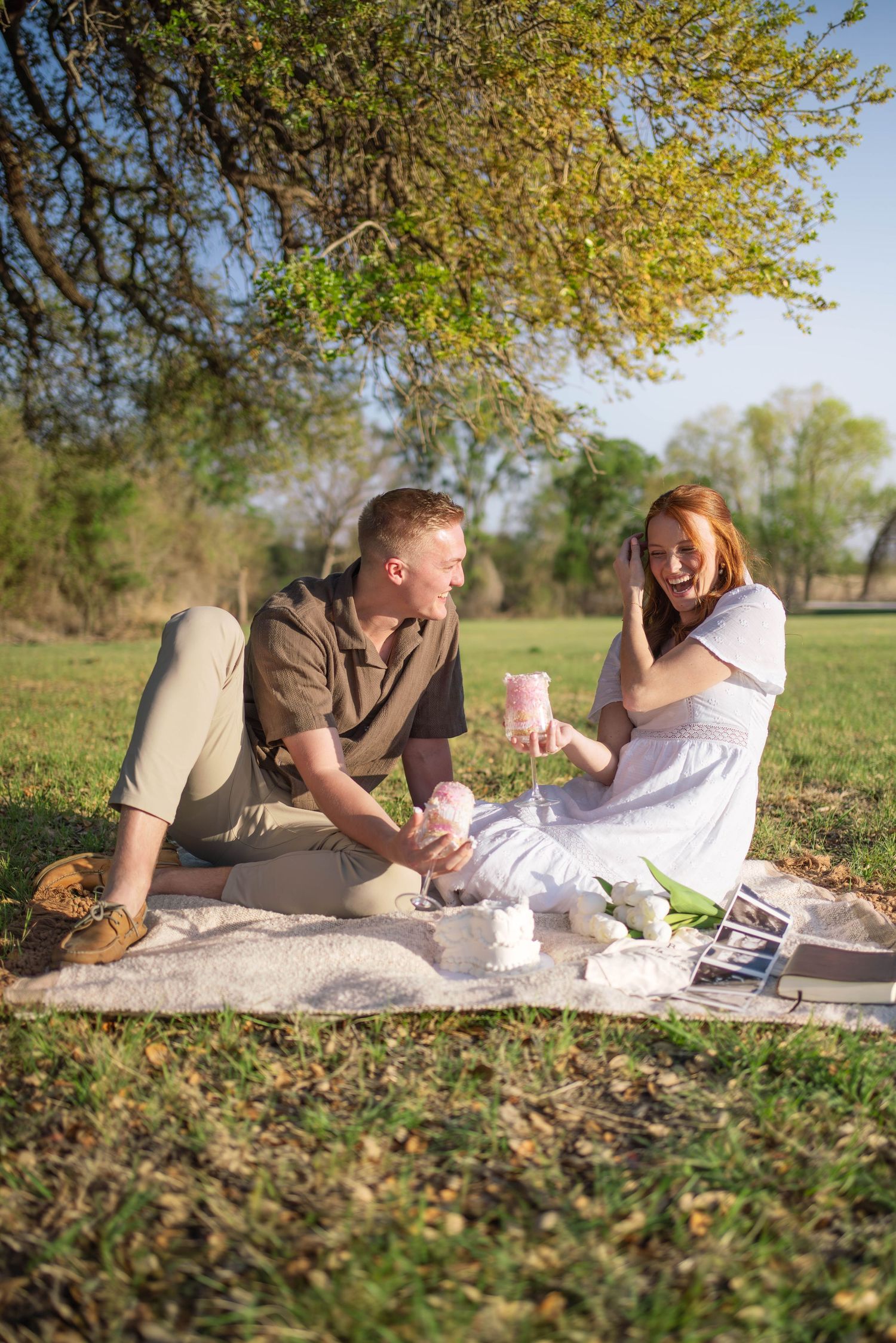 Happy couple sits on blanket in park sharing pink drinks during a sunny outdoor picnic.