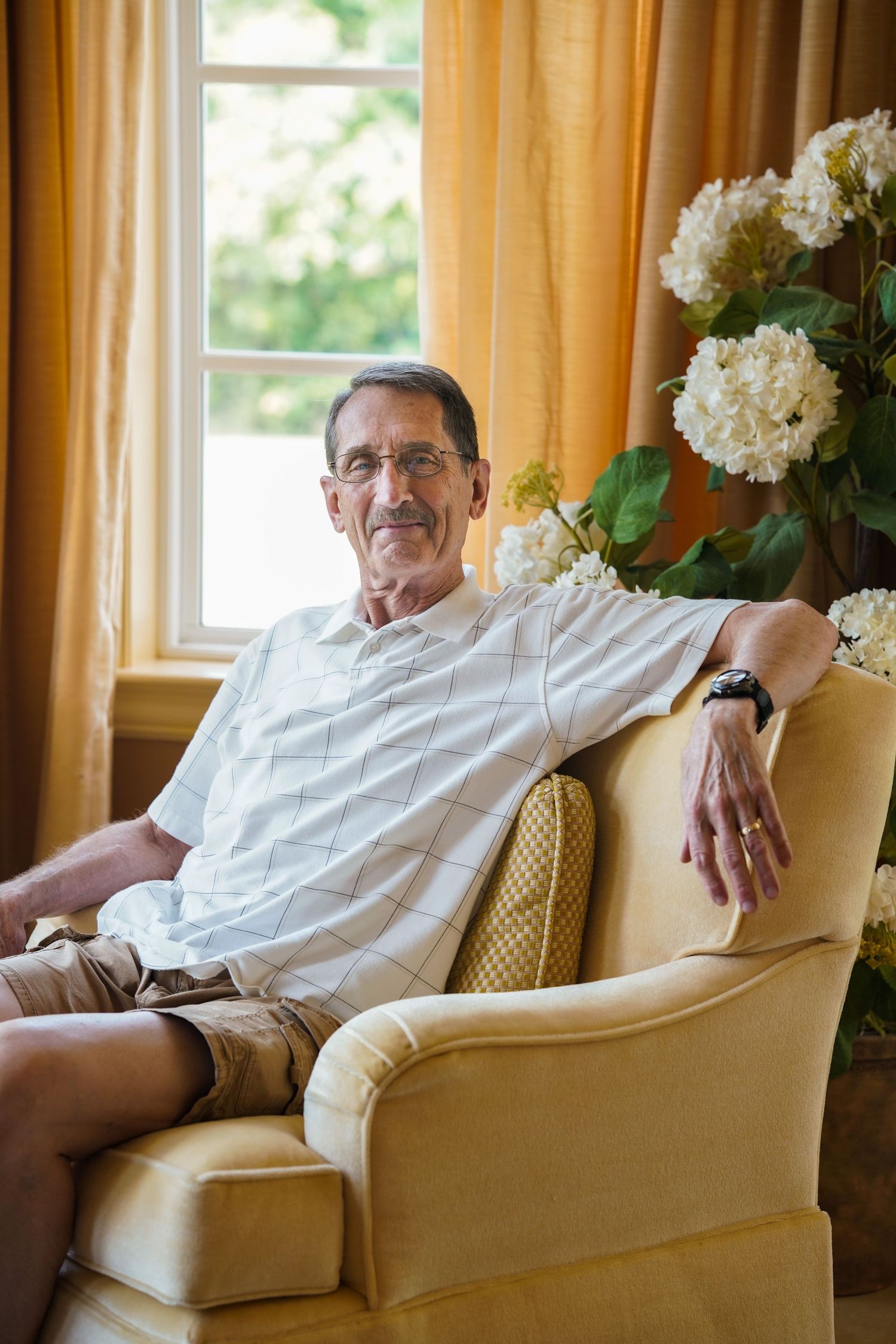 Elderly man in glasses and casual attire relaxing in a yellow armchair near a window with white flowers.