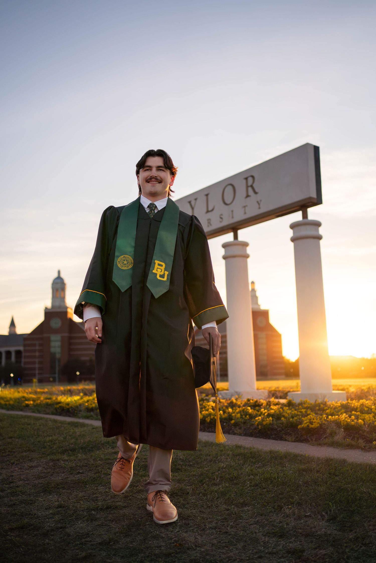 Baylor University graduate in green cap and gown poses proudly in front of campus sign at golden hour sunset.